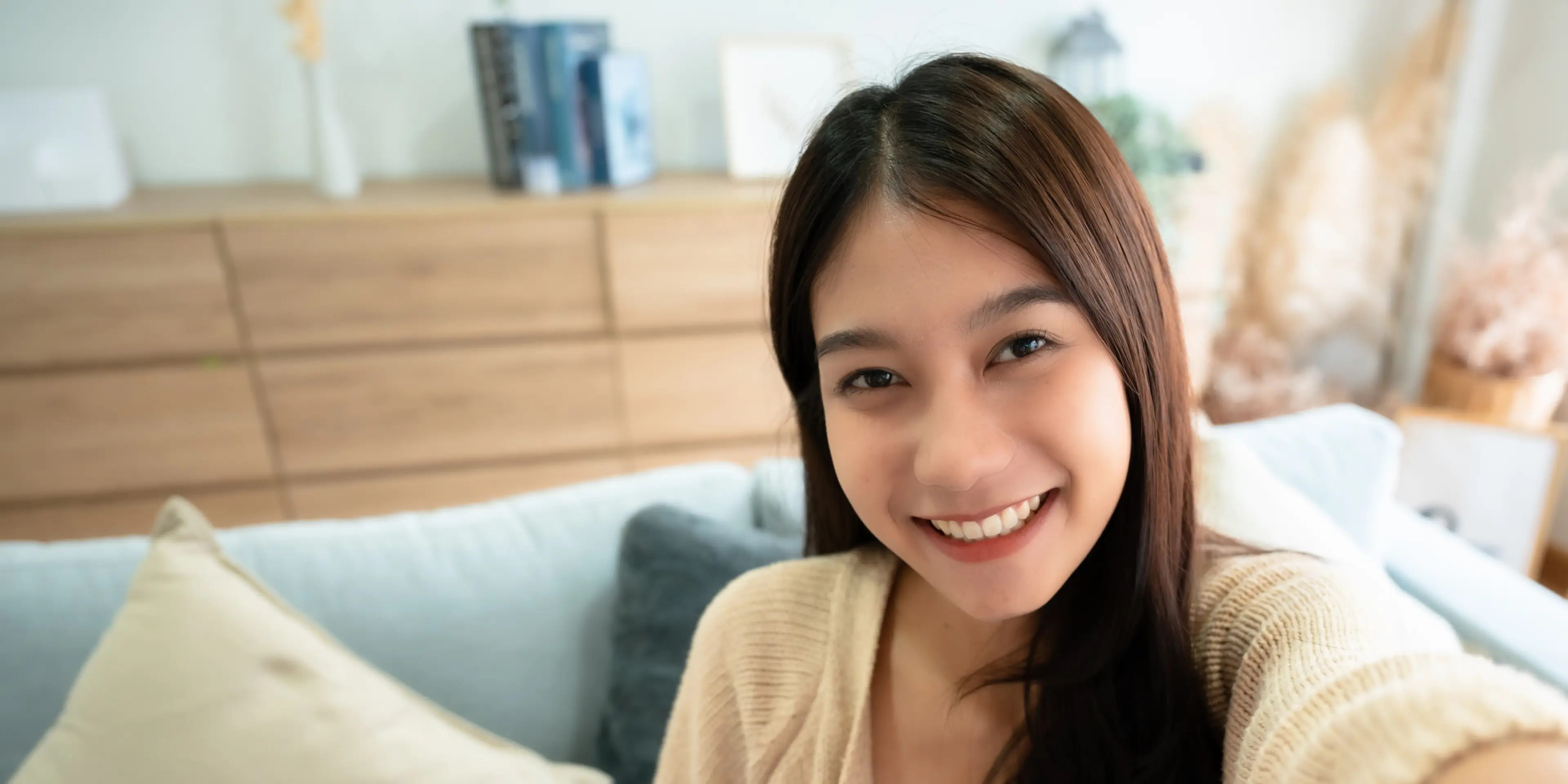Young woman with long dark hair smiling and wearing a beige sweater sitting on a sofa with cushions.