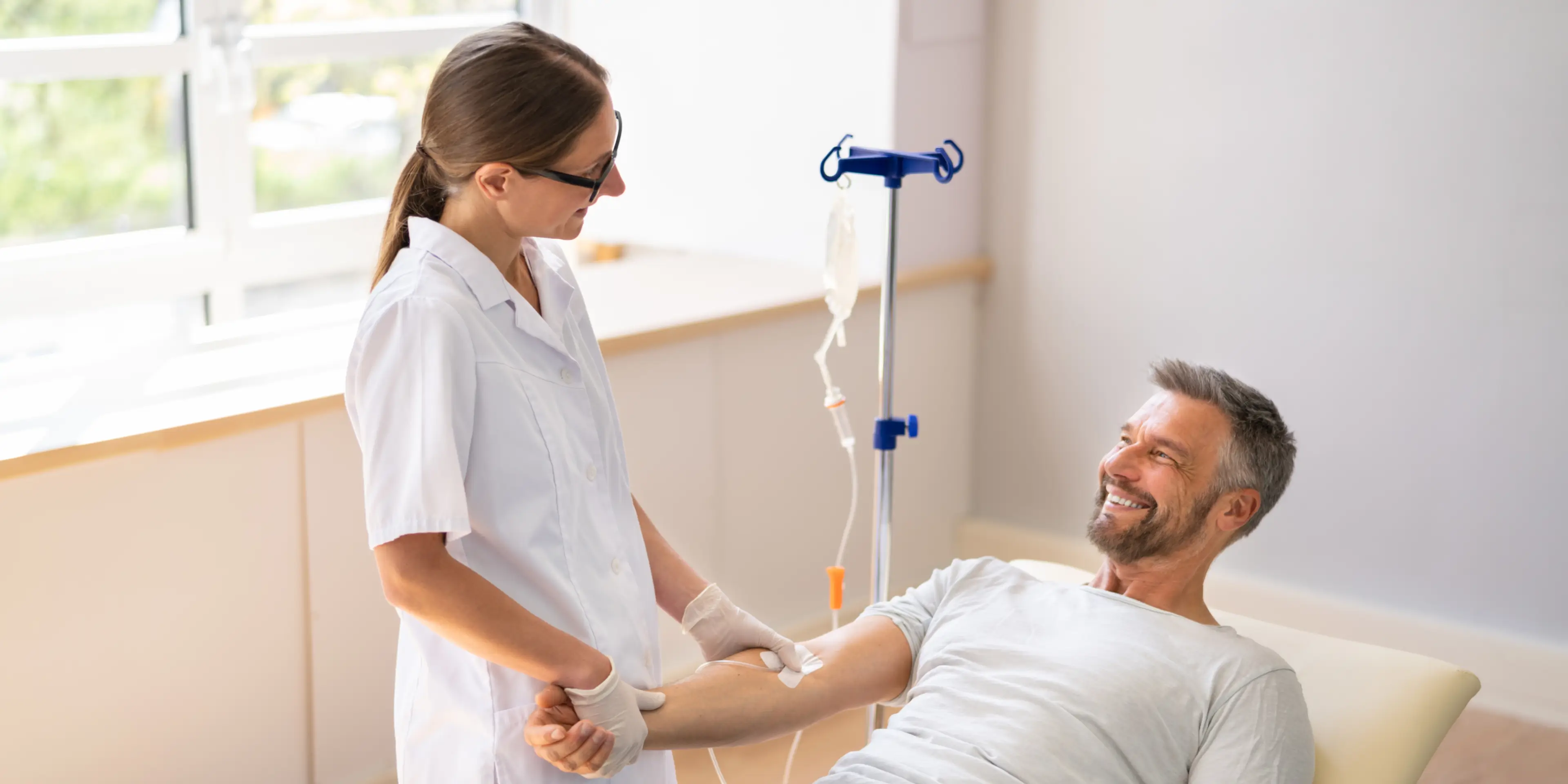 Smiling male patient receiving intravenous treatment from a female healthcare worker in a bright medical room.
