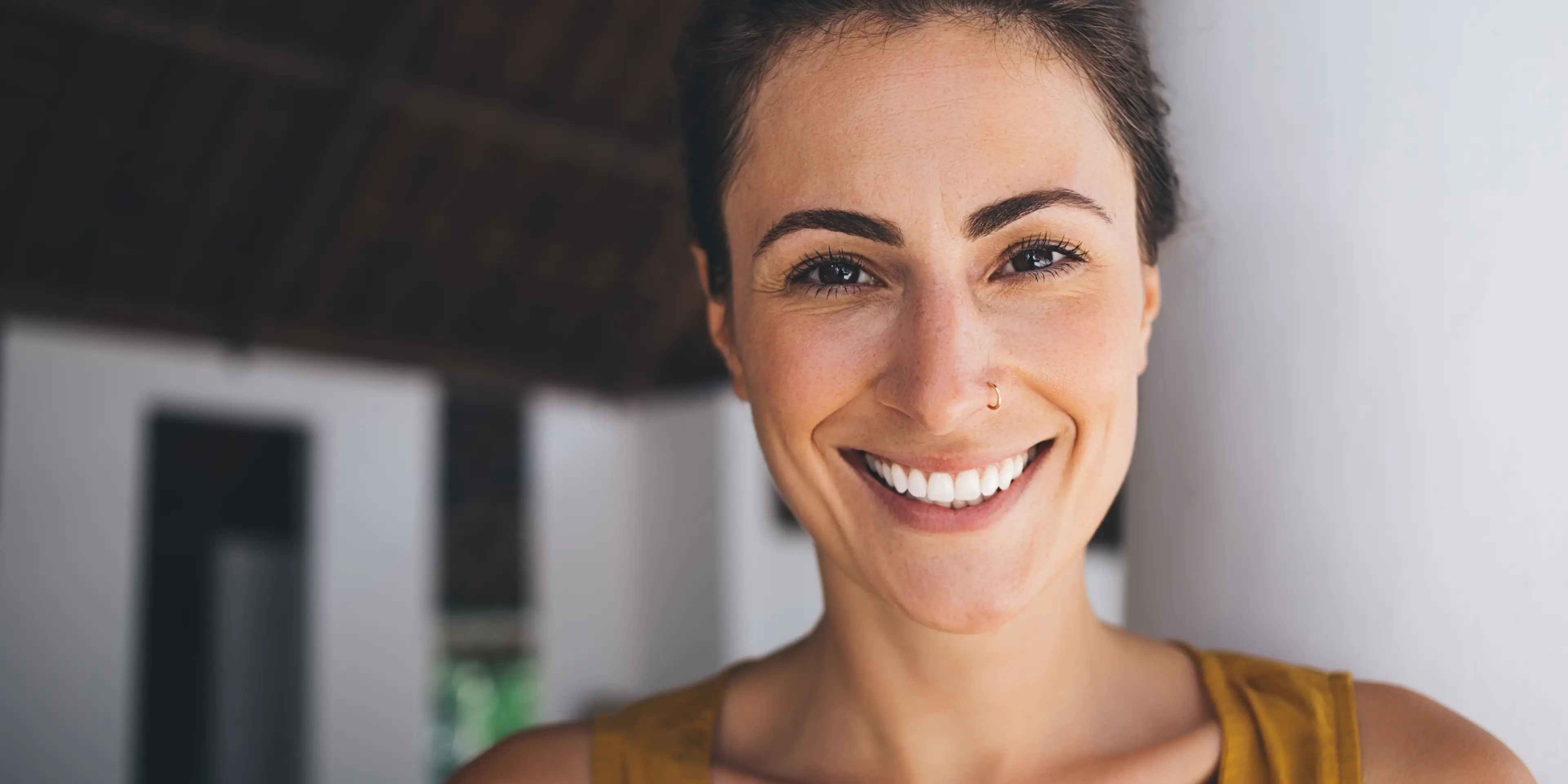Close-up of a smiling woman with a nose ring and dark hair, wearing a mustard yellow top, leaning against a white wall.