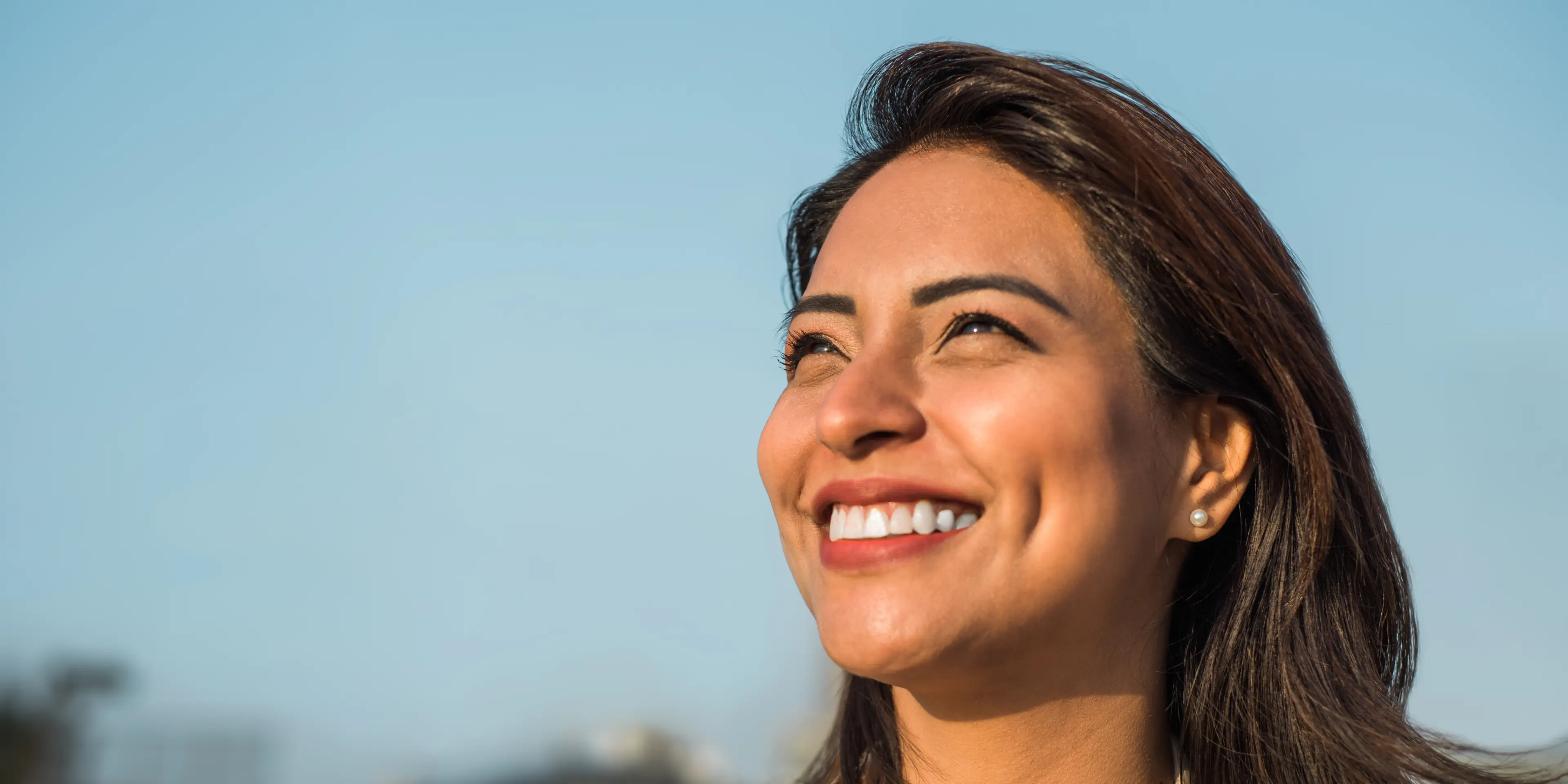 Close-up of a woman with dark hair smiling brightly against a clear blue sky.