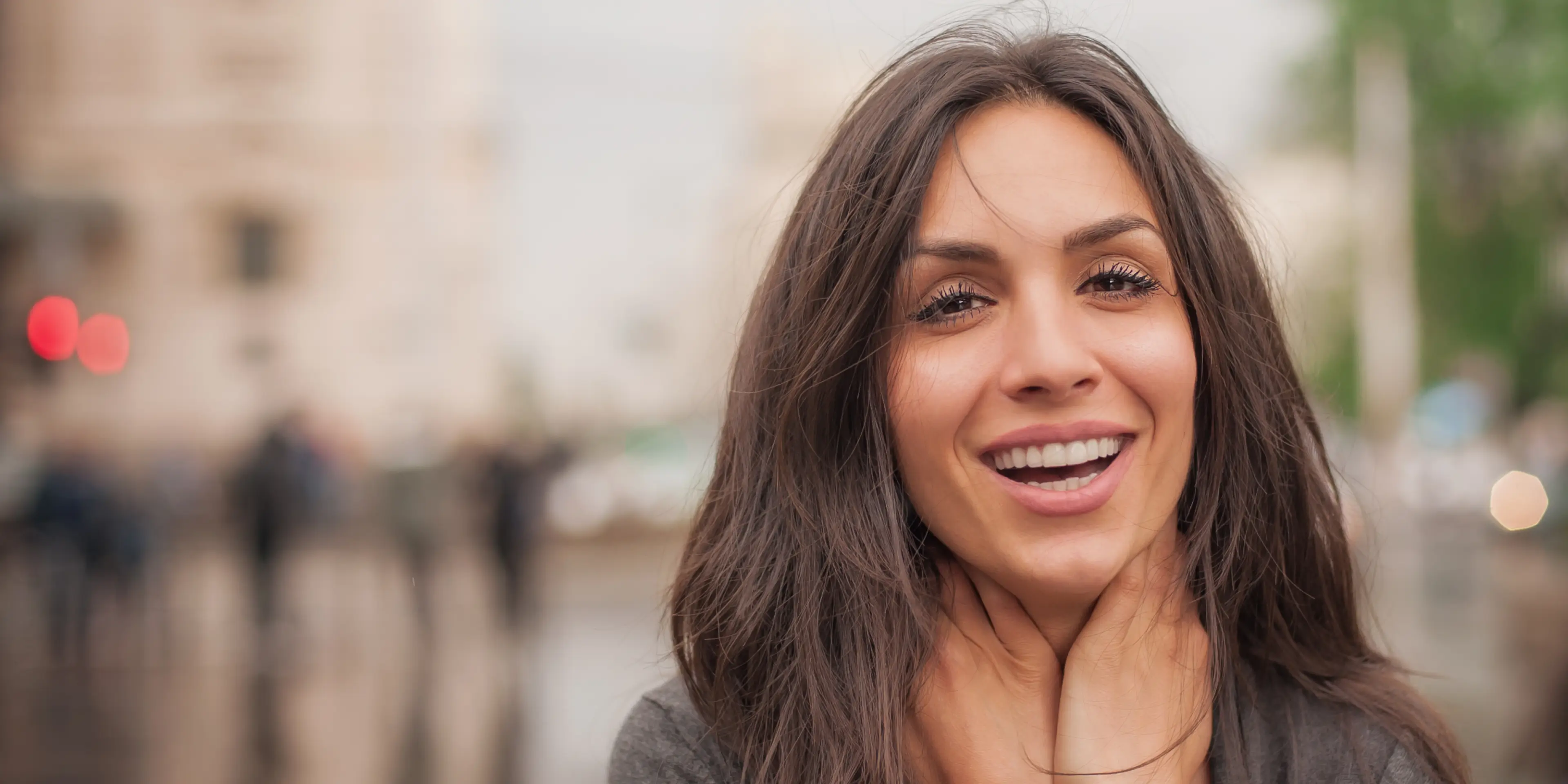 Close-up of a smiling woman with long brown hair holding her neck outdoors with a blurred urban background.