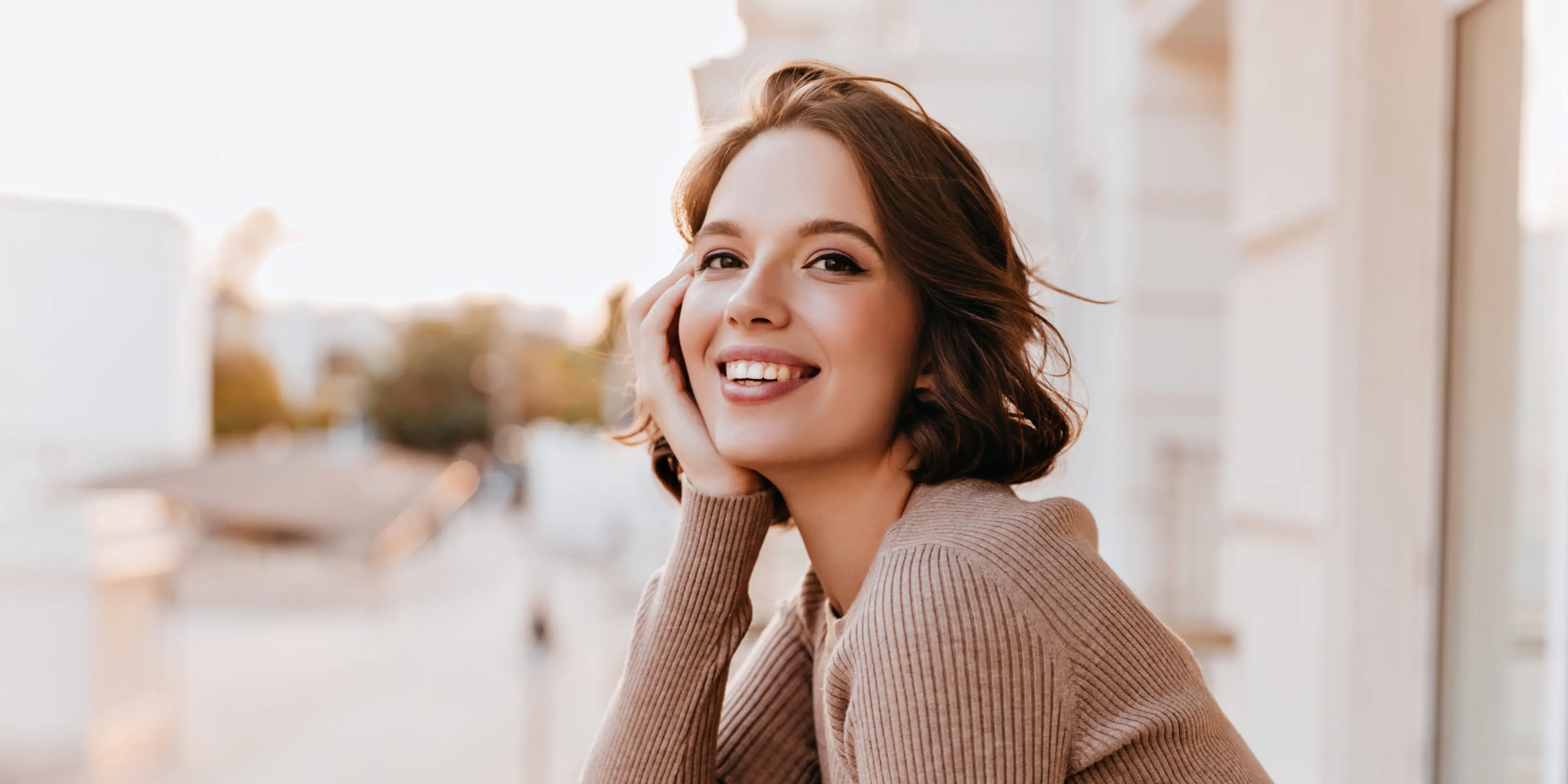 Young woman with short brown hair smiling and resting her face on her hand outdoors.