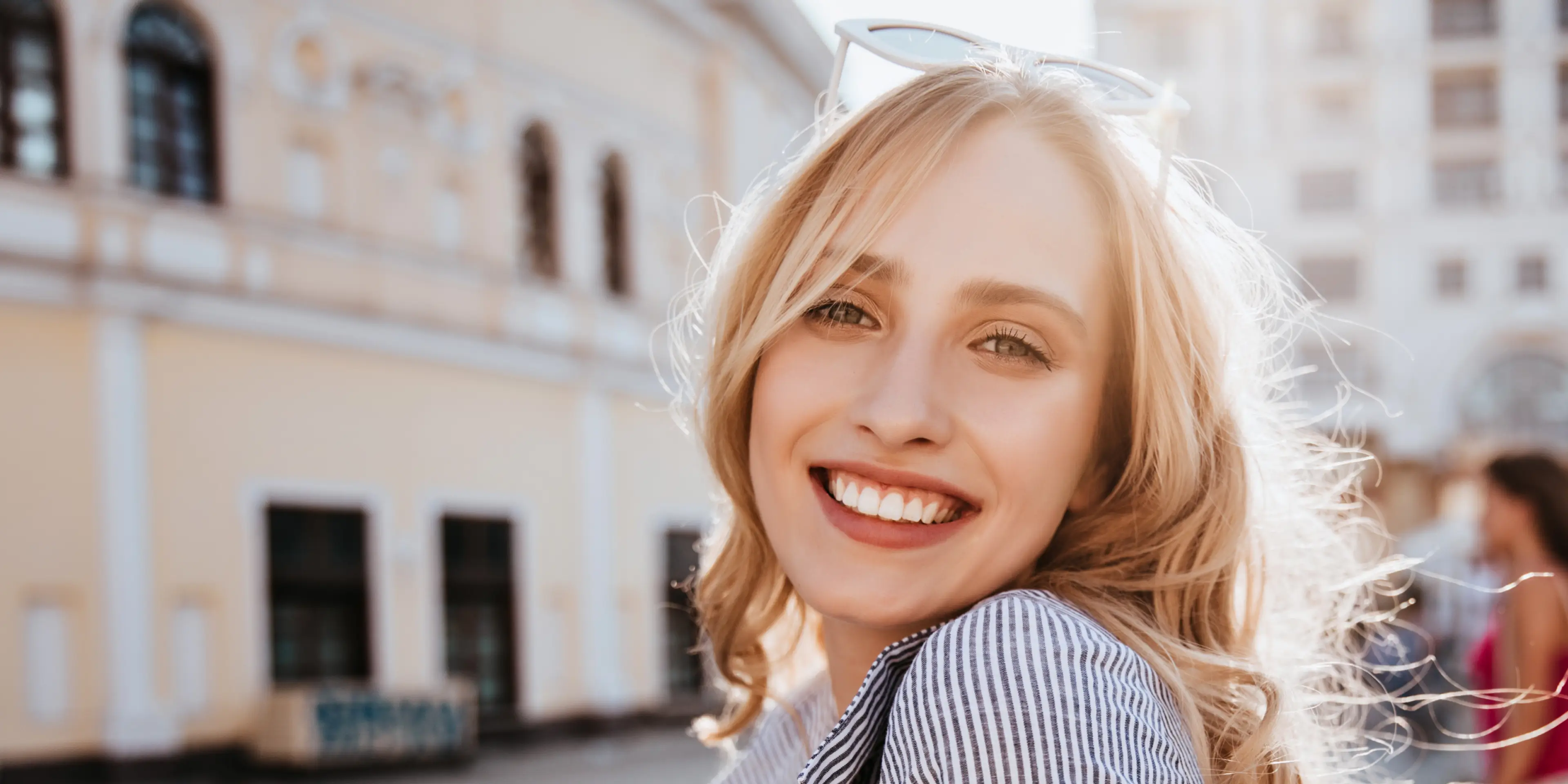 Smiling blonde woman outdoors with sunglasses on her head and wavy hair illuminated by sunlight.