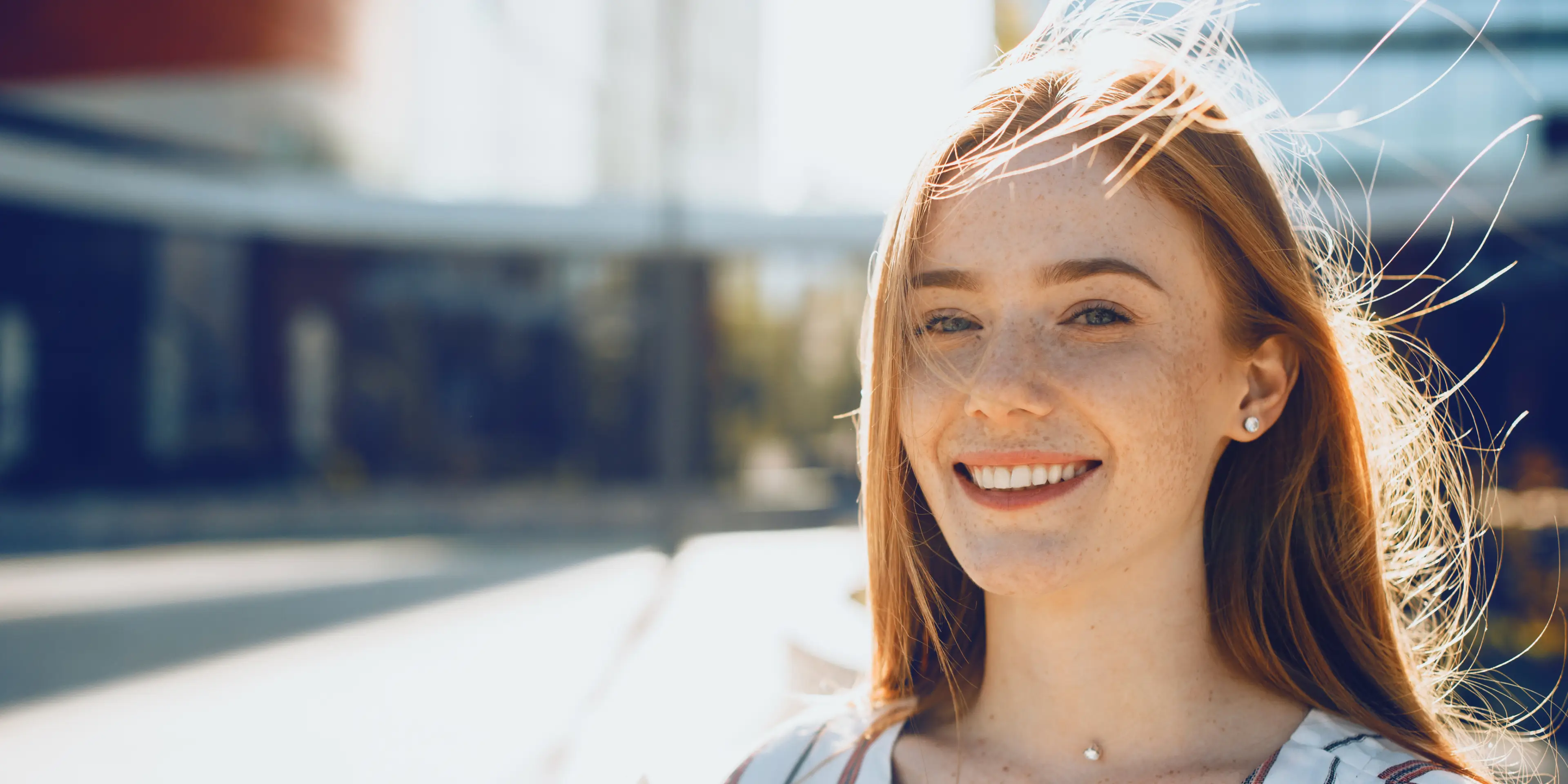 Smiling young woman with long red hair and freckles, standing outdoors in bright sunlight.