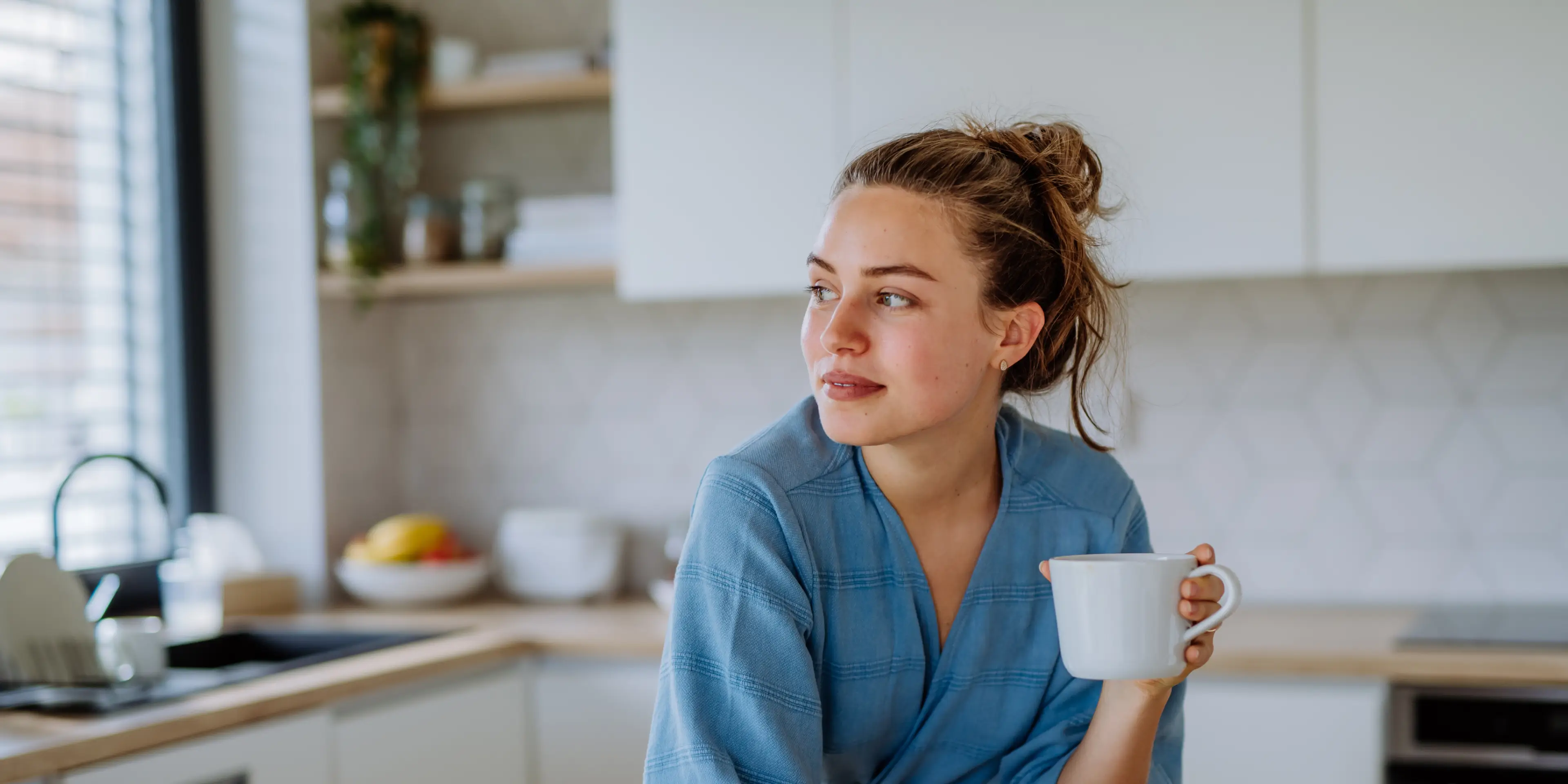 Young woman in a blue robe holding a white mug and looking to the side in a bright modern kitchen.