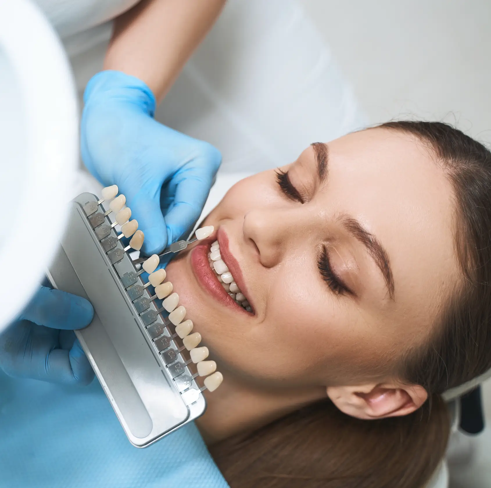 Dentist with blue gloves matching a dental veneer shade to a smiling woman's teeth.
