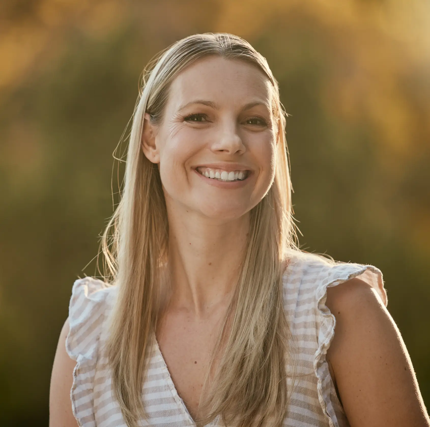 Smiling woman with long blonde hair wearing a striped sleeveless top, with a blurred nature background.