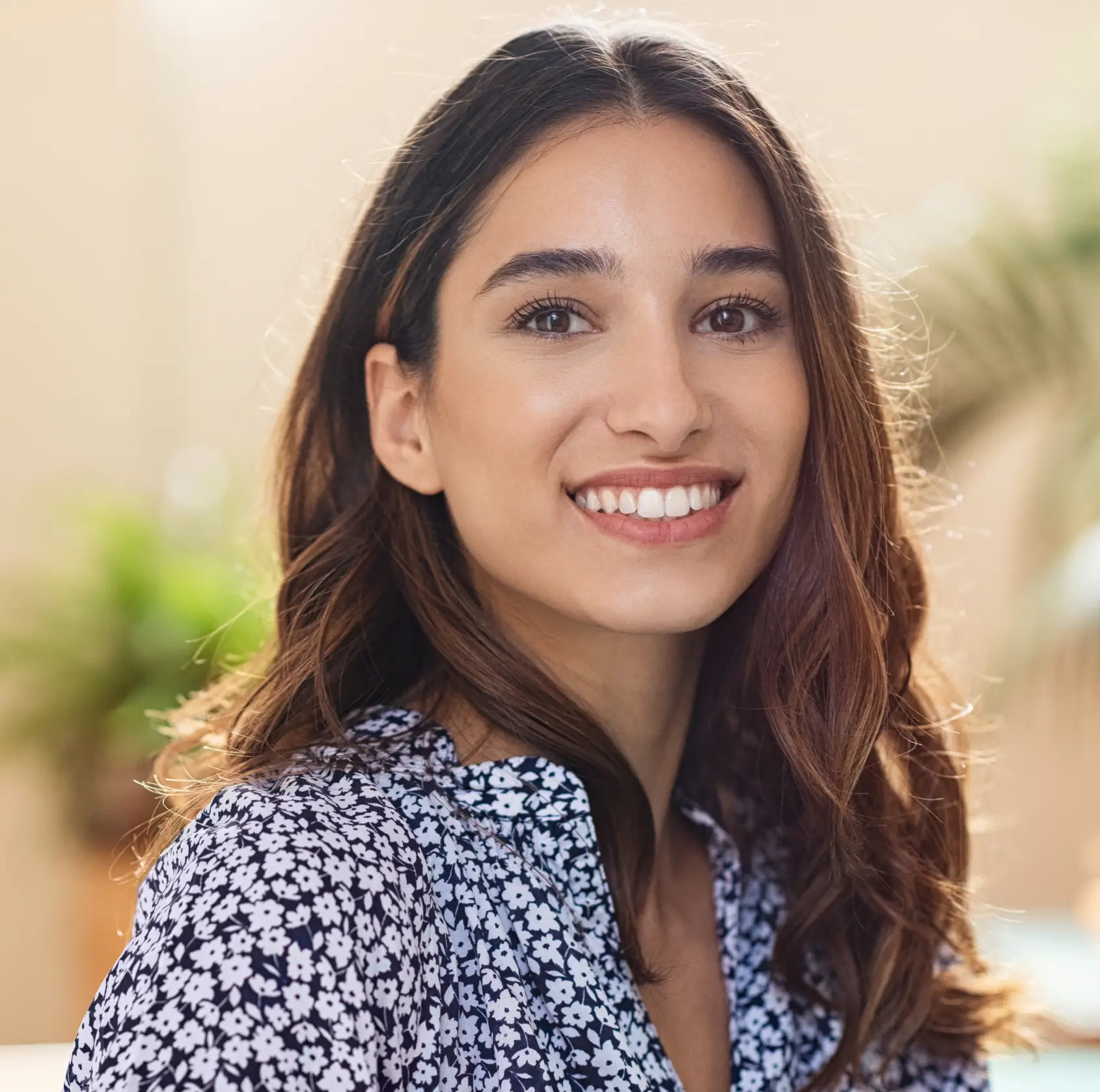 Smiling woman with long brown hair wearing a black and white floral blouse in soft natural light.