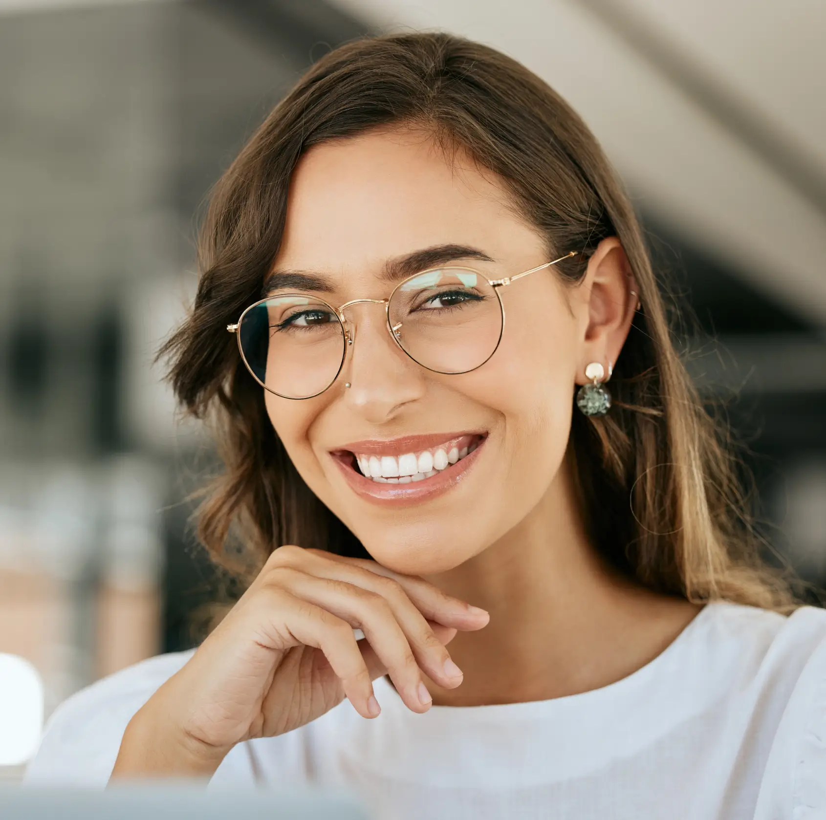 Smiling woman with glasses, brown hair, and earrings, resting her chin on her hand.