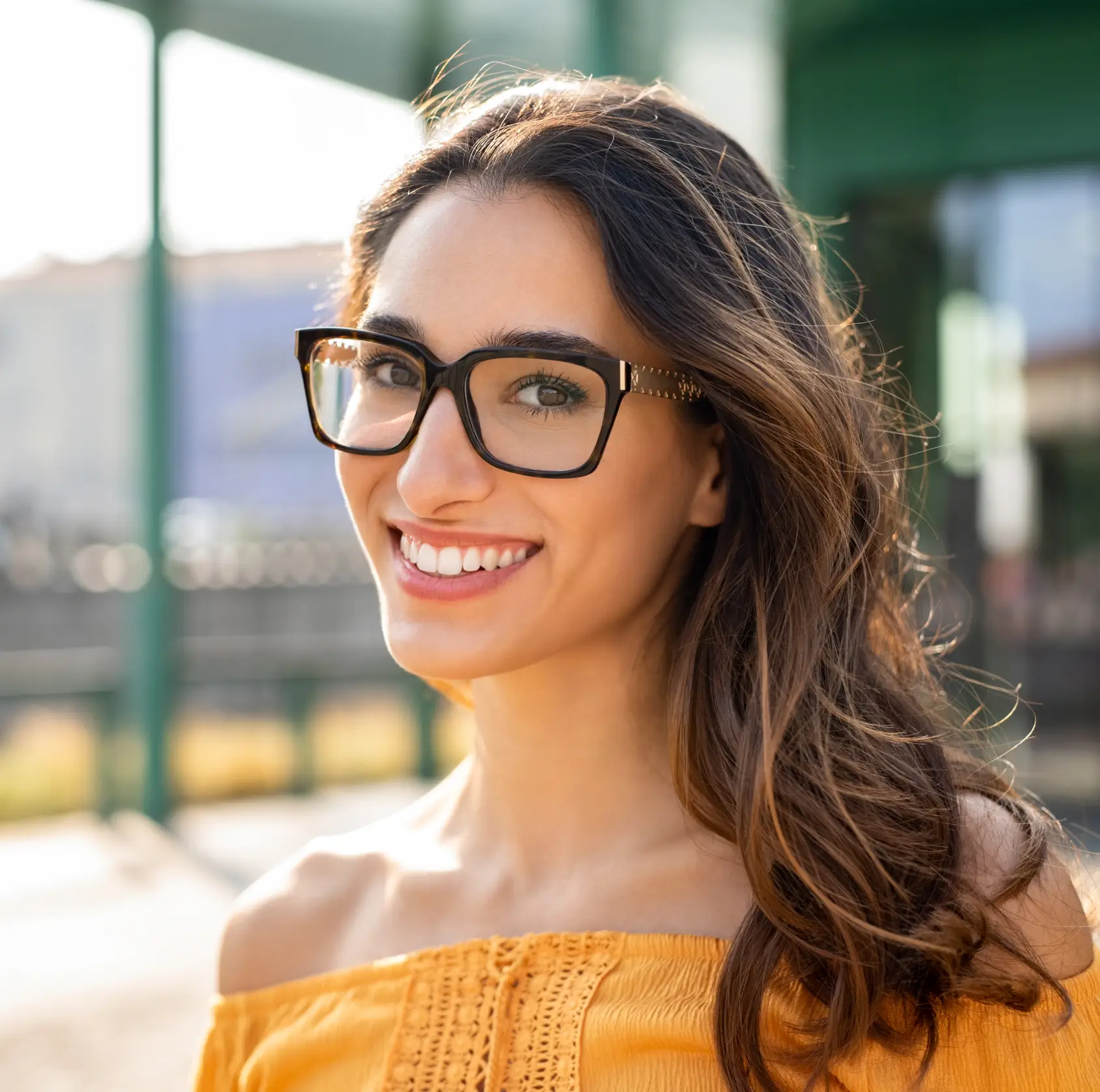 Smiling woman with long brown hair wearing black glasses and an off-shoulder yellow top outdoors.