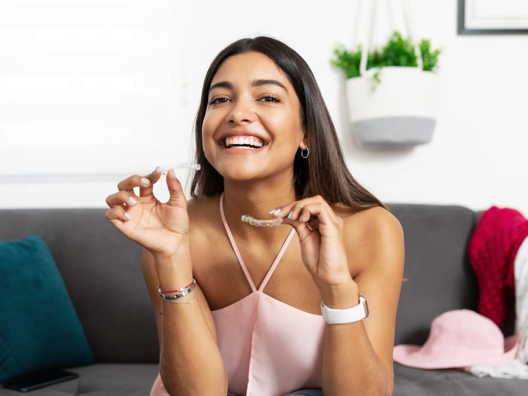 Smiling woman sitting on couch holding clear dental aligners in both hands.