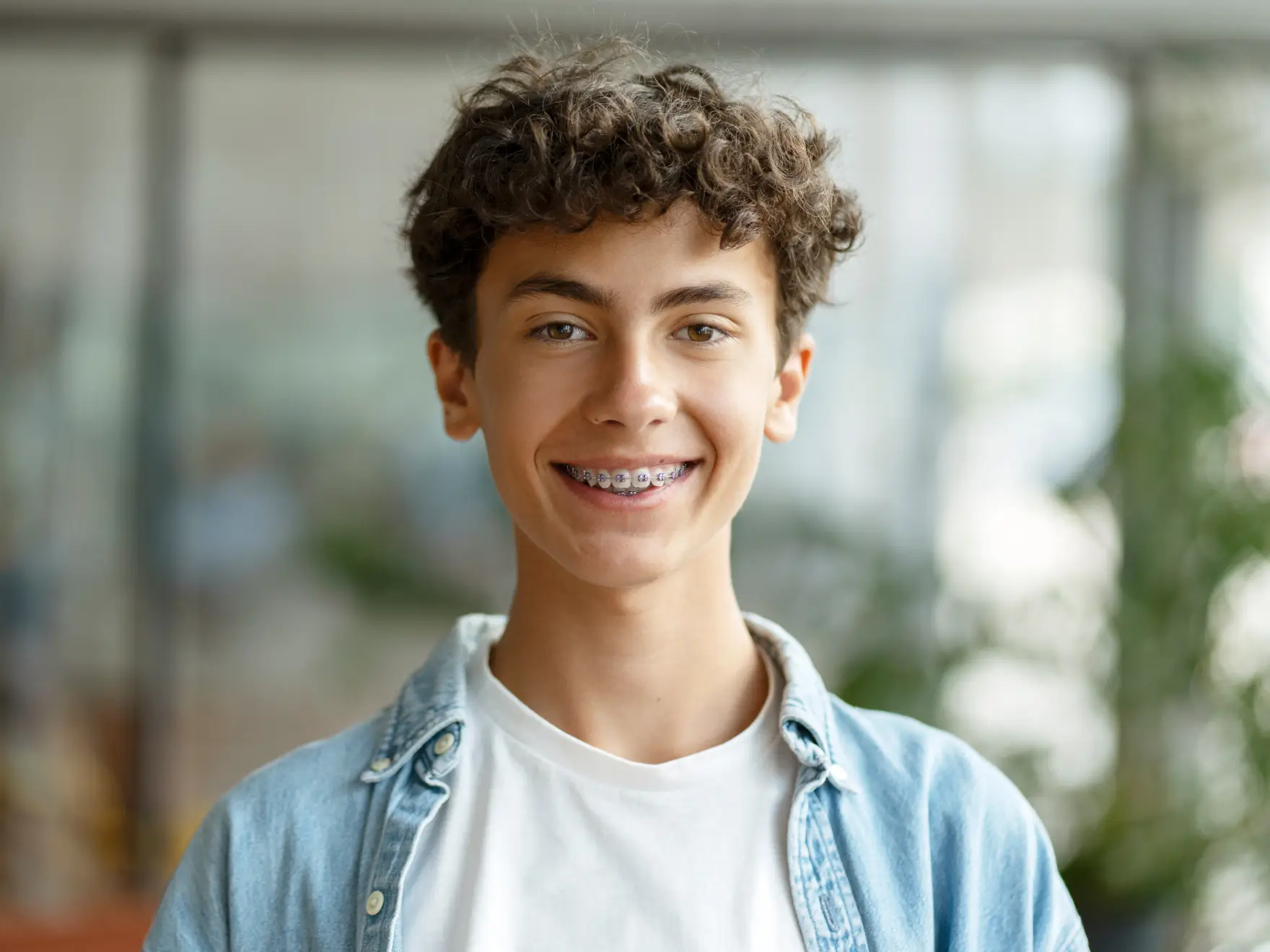 Smiling teenage boy with curly hair wearing braces, a white t-shirt, and a light denim shirt.