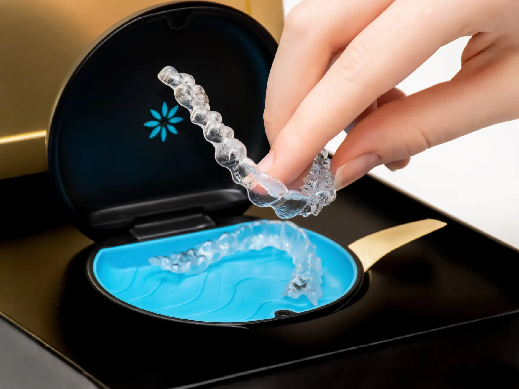 Hand holding a clear dental aligner above an open black and blue aligner storage case.