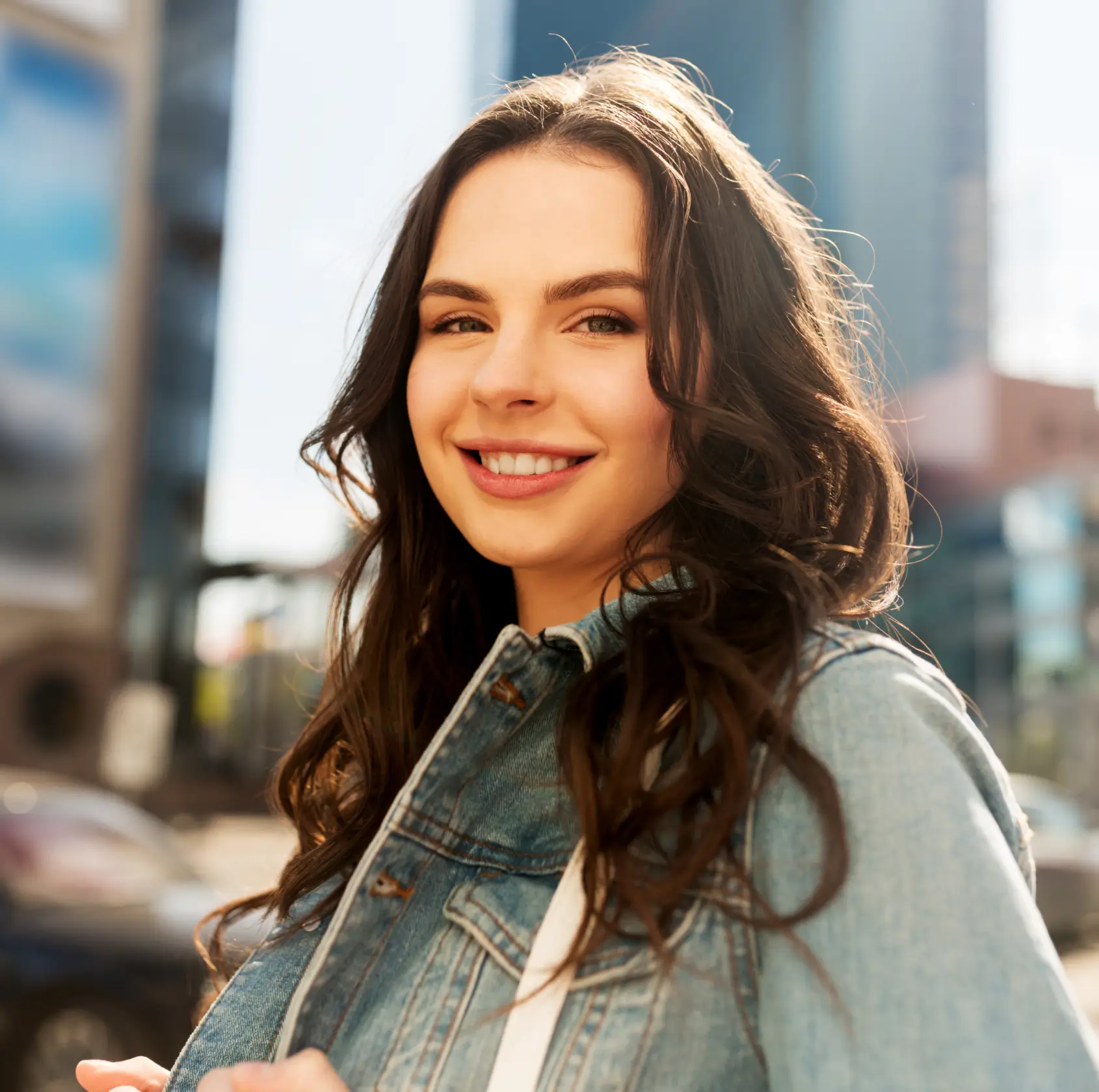 Smiling young woman with long brown hair wearing a denim jacket in an urban setting.