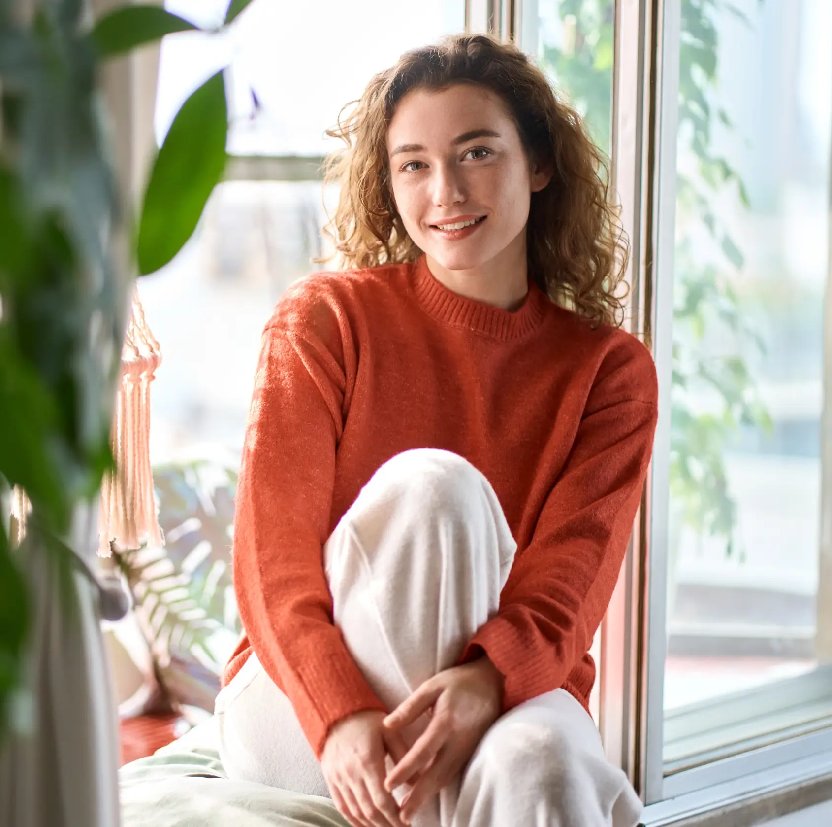 Smiling woman with curly hair wearing an orange sweater and white pants sitting by a window indoors.