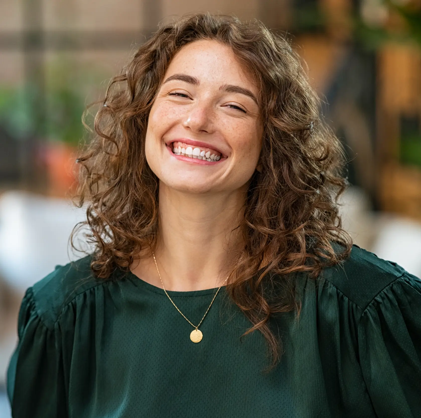 Smiling young woman with curly brown hair wearing a dark green top and a gold pendant necklace.