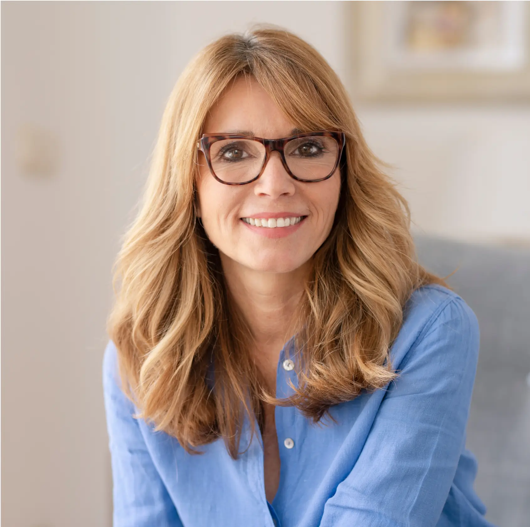 Smiling middle-aged woman with long blonde hair and glasses wearing a blue shirt, sitting indoors.