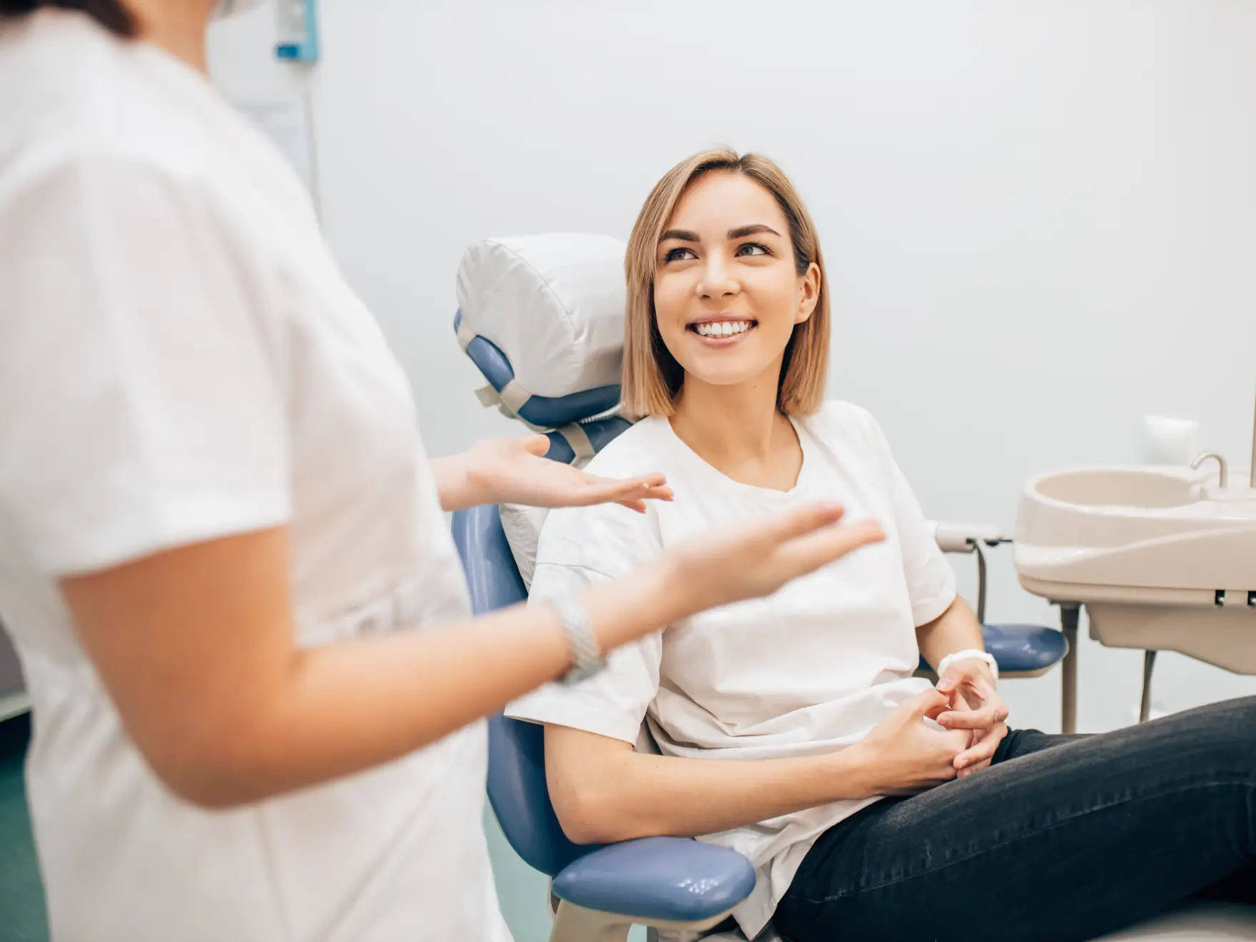 Smiling woman sitting in a dental chair talking to a healthcare professional in white uniform.