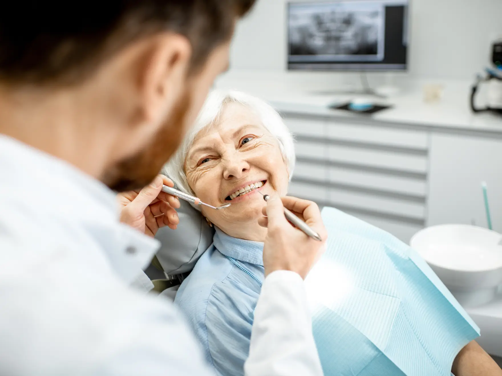 Elderly woman smiling during a dental checkup with a dentist holding dental instruments near her mouth.