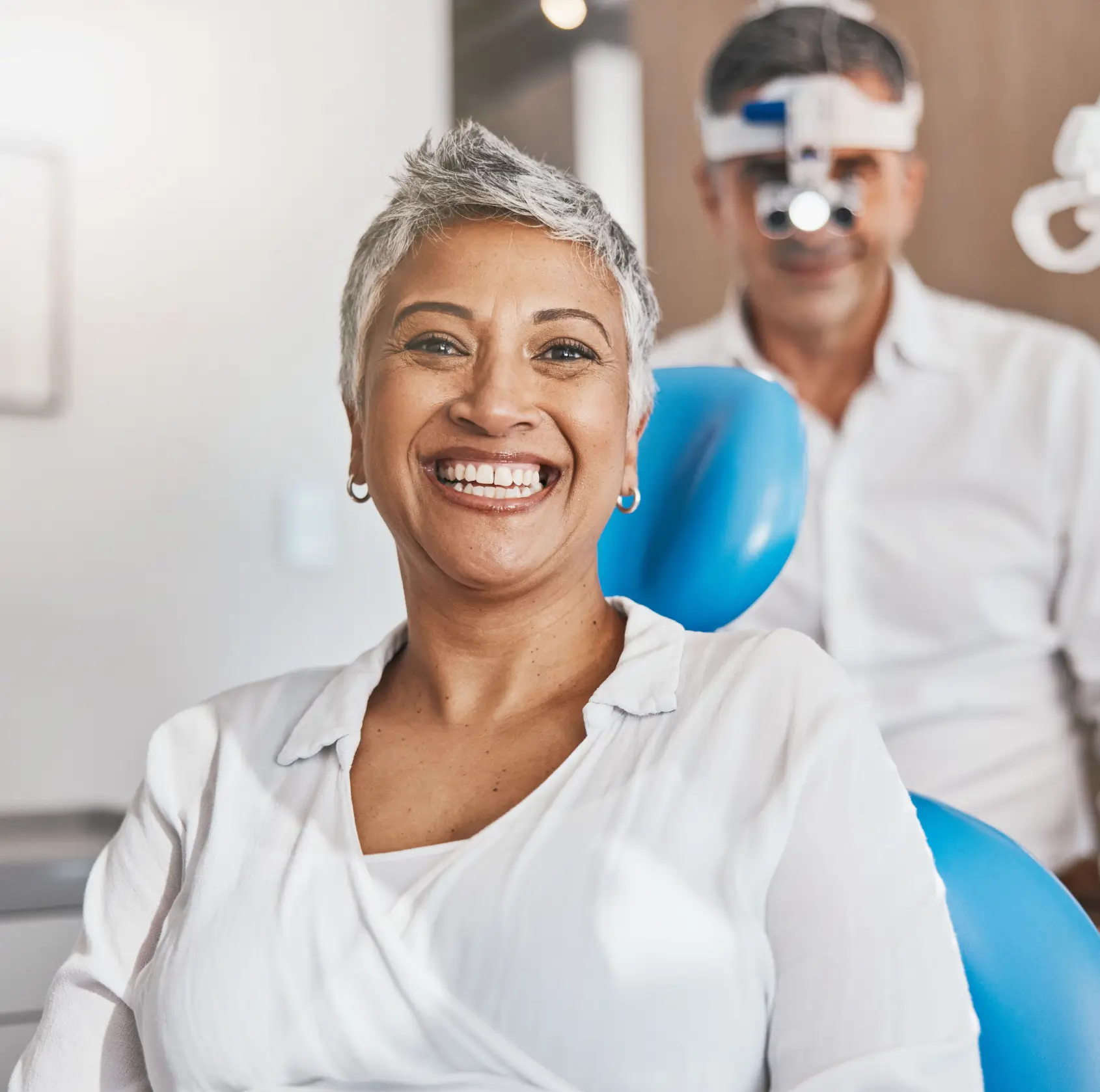 Smiling middle-aged woman sitting in a dental chair with a dentist wearing magnifying glasses in the background.