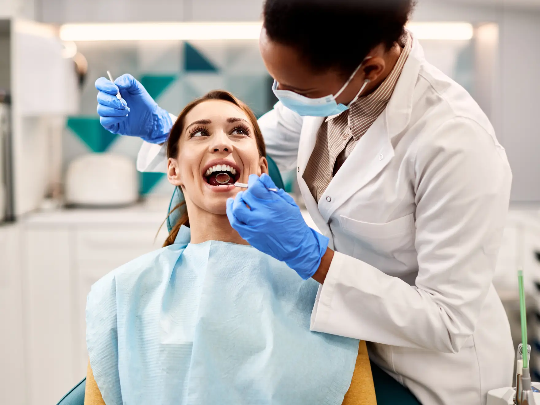 Dentist wearing gloves and a mask examining a smiling female patient's teeth with dental tools.