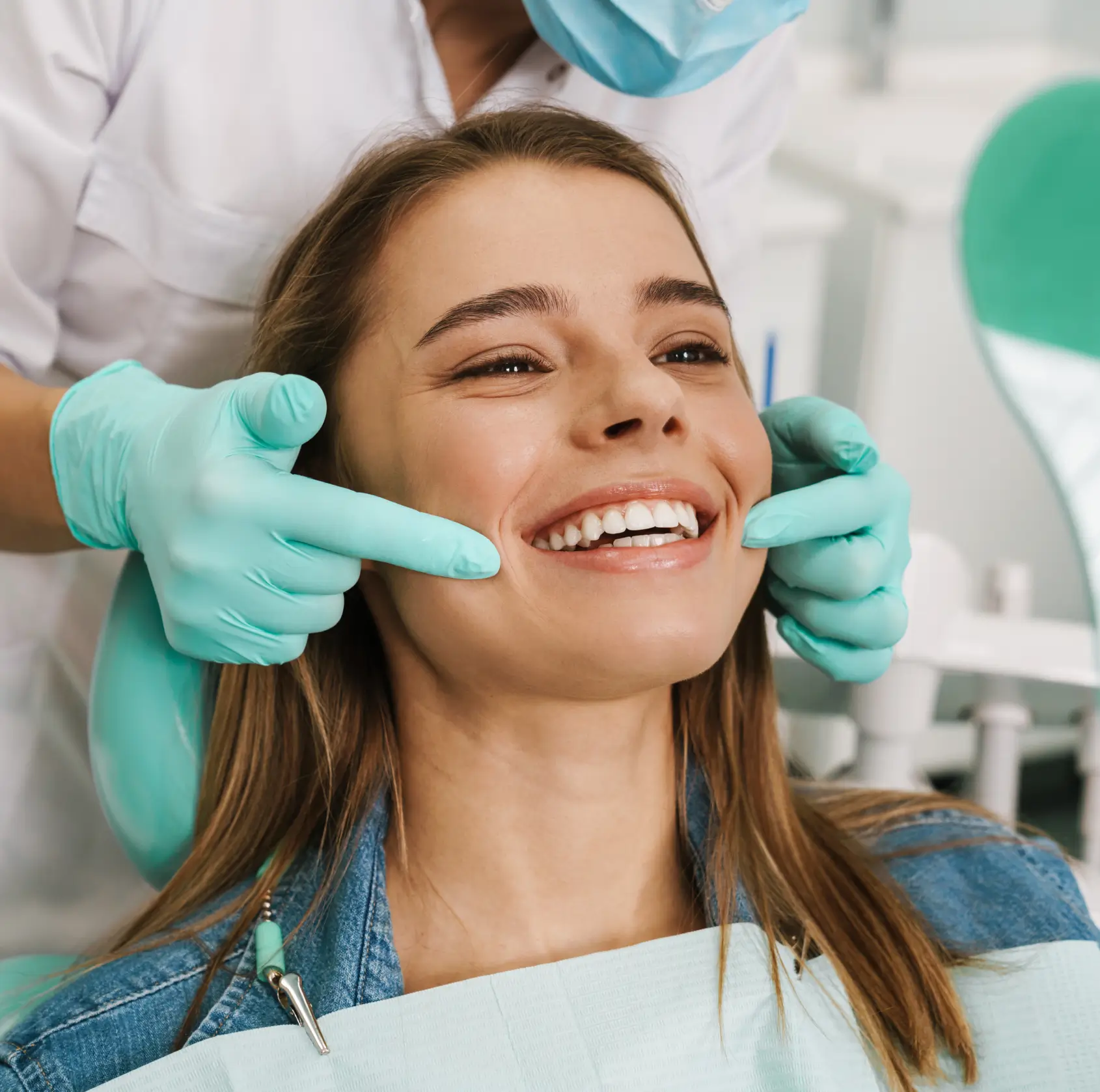 Dentist wearing gloves and a mask examines a smiling young woman's teeth in a dental clinic.