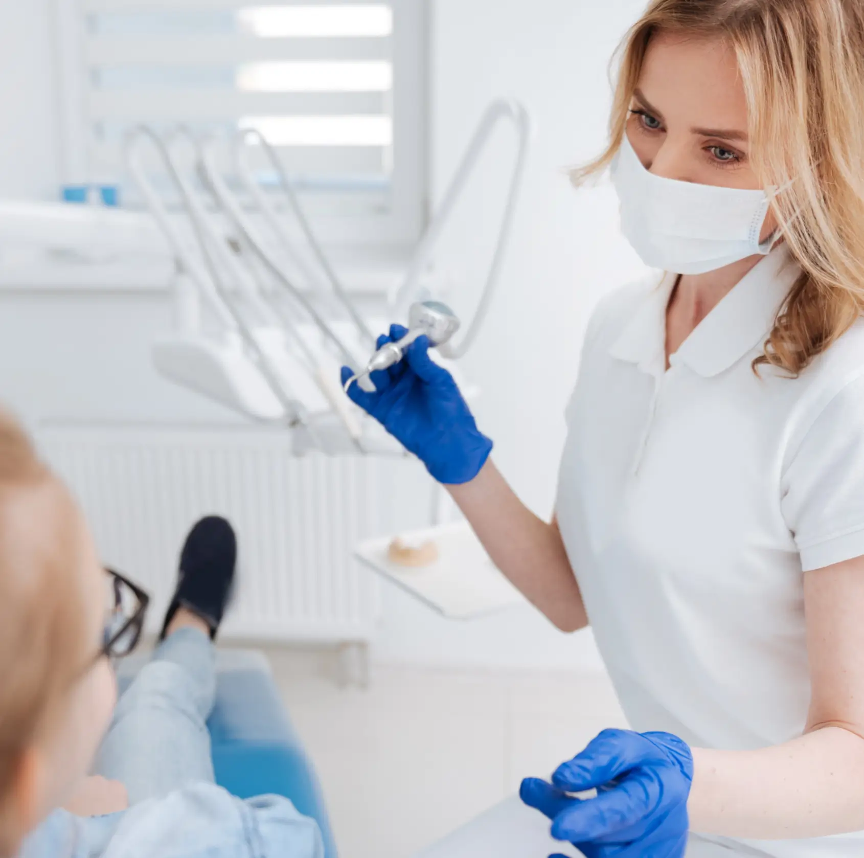 Dentist wearing a mask and blue gloves holding a dental tool while attending to a patient in a dental clinic.