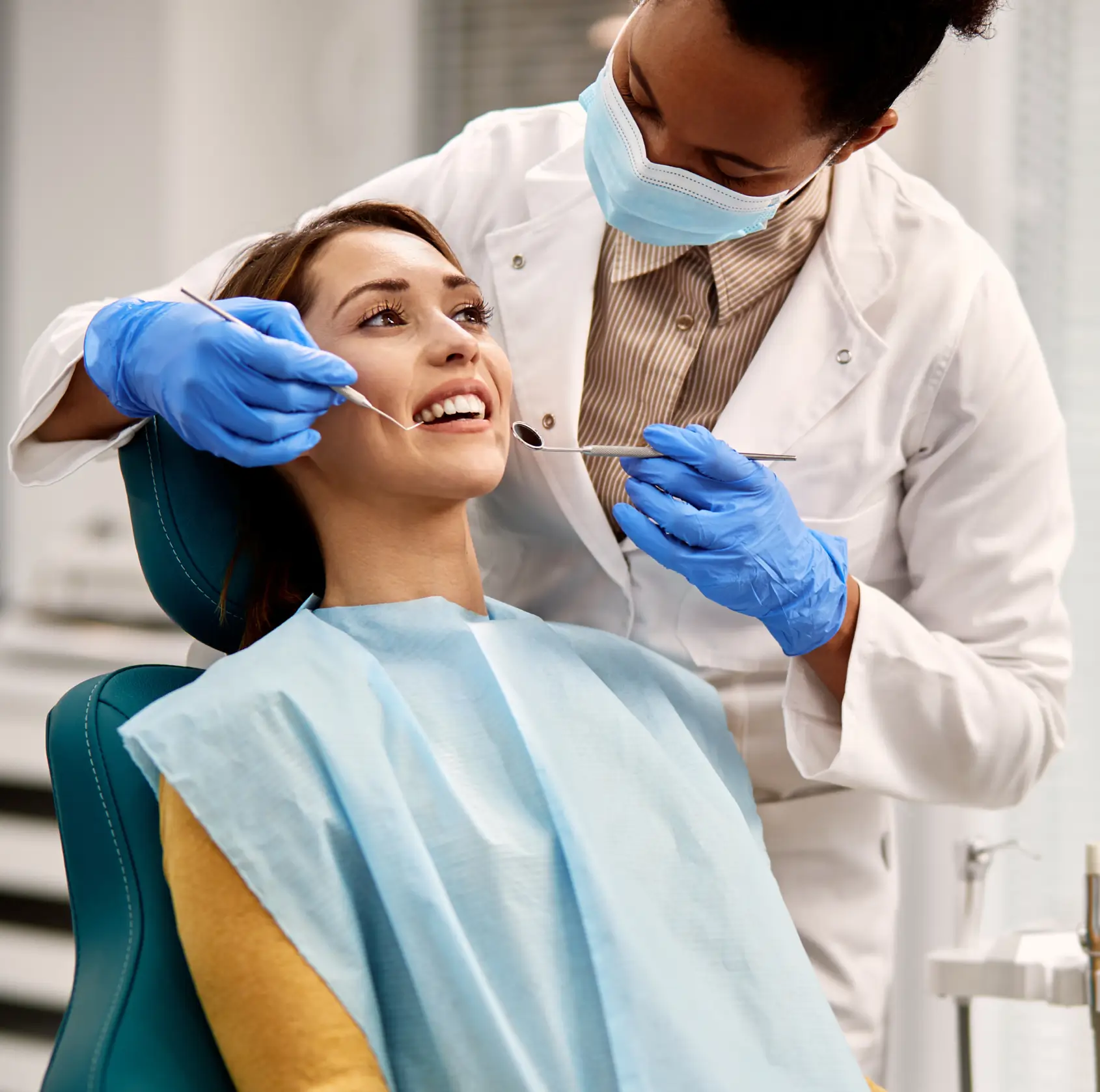 Dentist wearing mask and gloves examining smiling patient's teeth with dental tools in clinic.