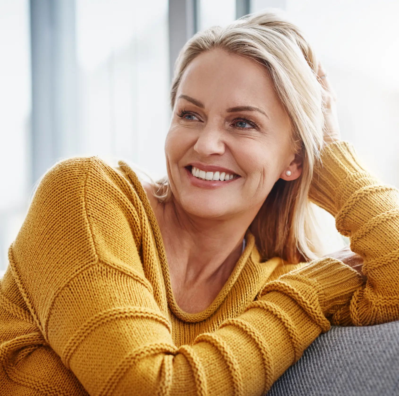 Smiling middle-aged woman with blonde hair wearing a mustard yellow knit sweater, leaning on a gray surface.