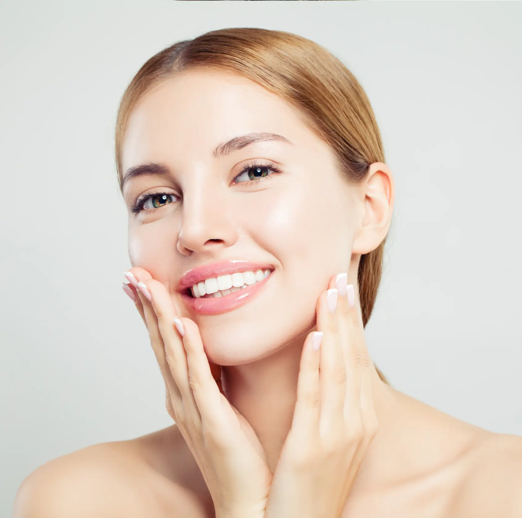 Smiling woman with clear skin gently touching her face against a light gray background.