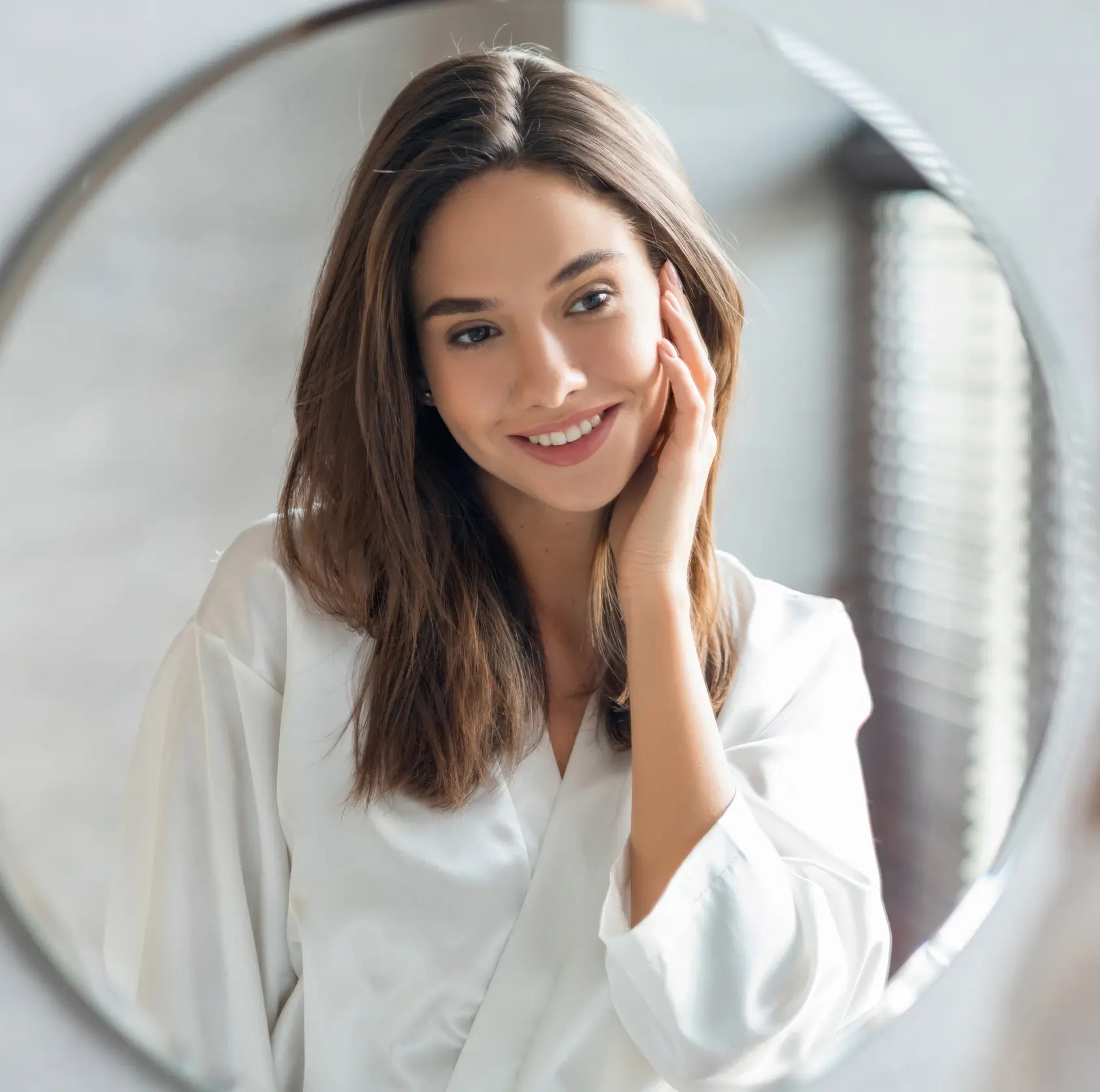 Woman with shoulder-length brown hair, wearing a white robe, smiling and looking at herself in a round mirror.