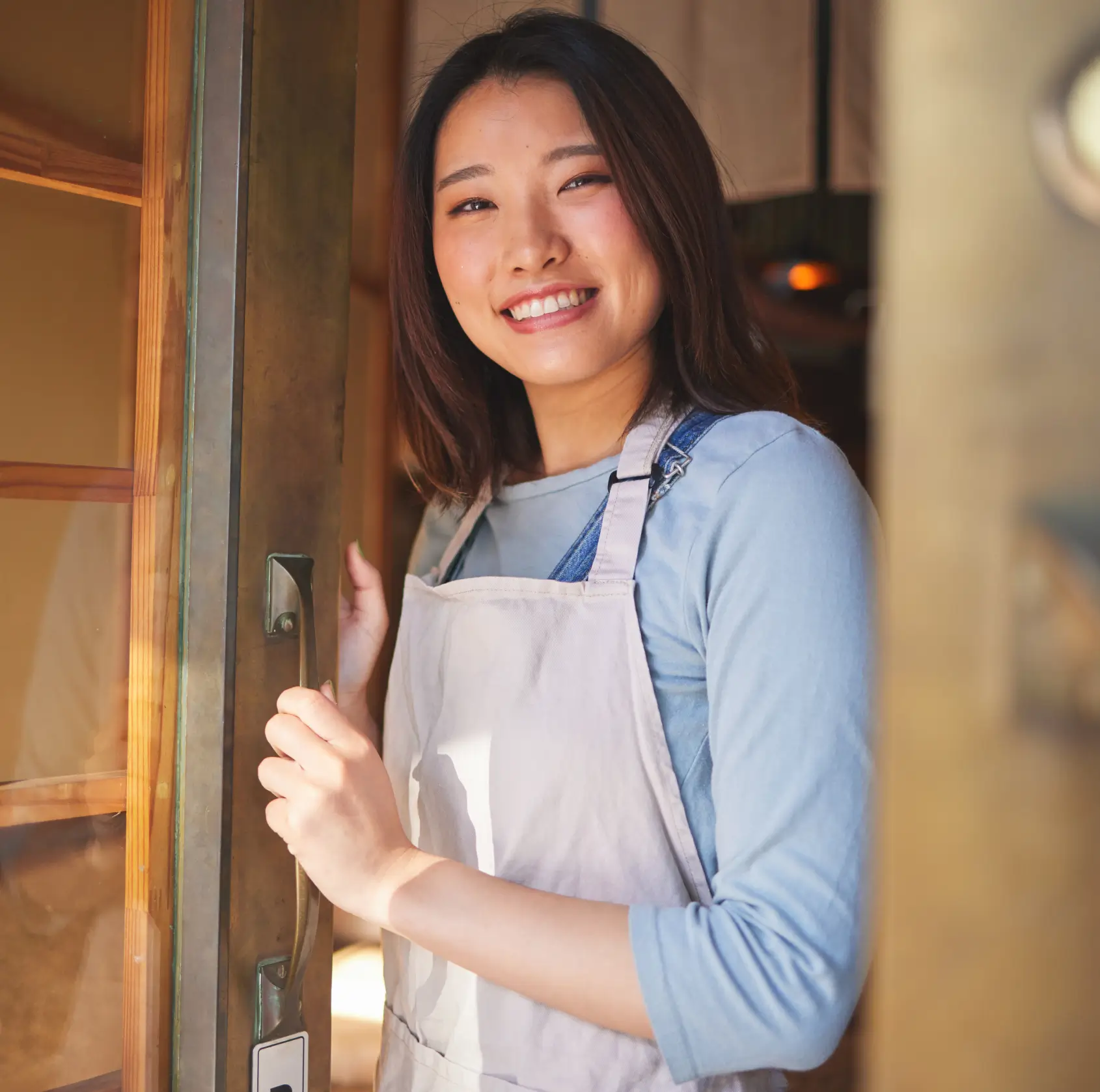 Smiling woman in apron opening a glass door and looking at the camera.