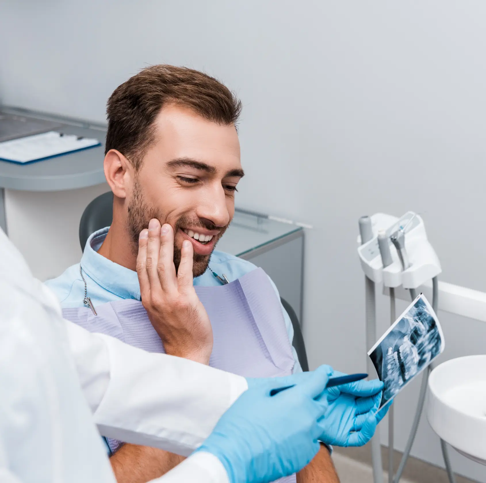 Smiling man at dental office holding his jaw while dentist shows him a dental X-ray.
