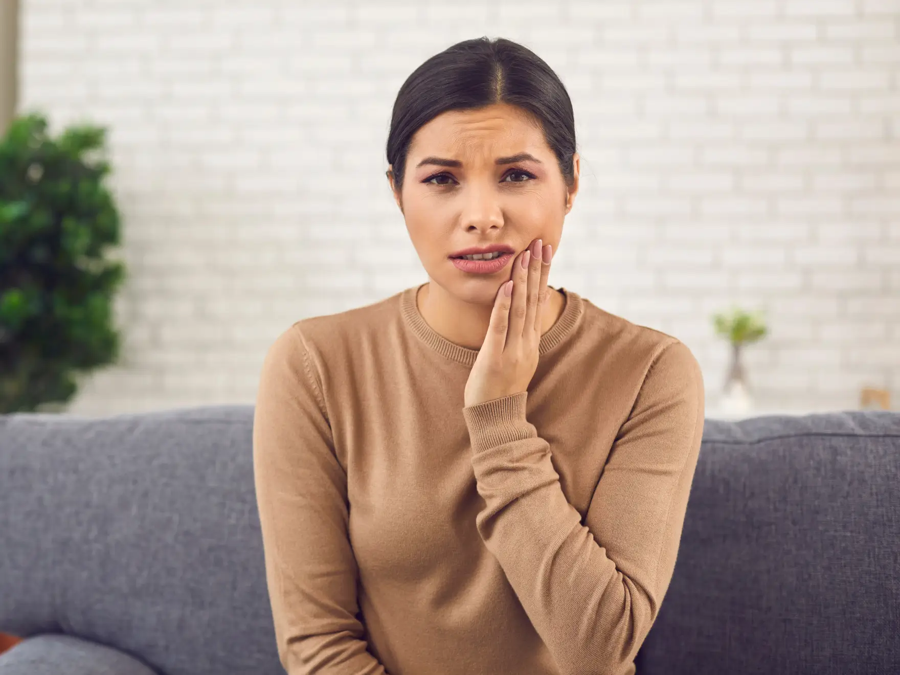 Woman sitting on a couch holding her cheek with a pained expression, suggesting toothache or jaw pain.