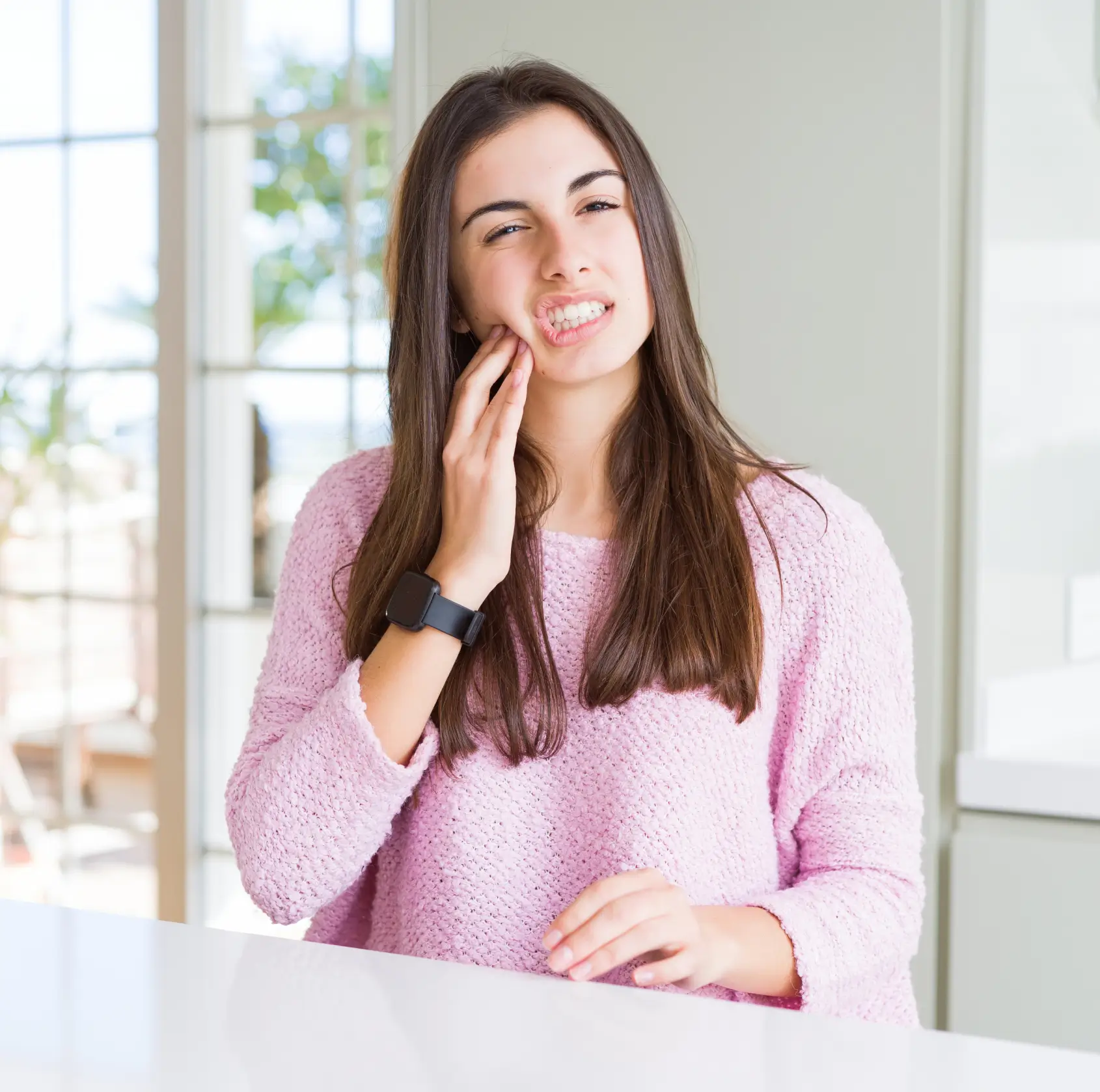 A young woman in a pink sweater touching her cheek, showing discomfort, indoors near a window.