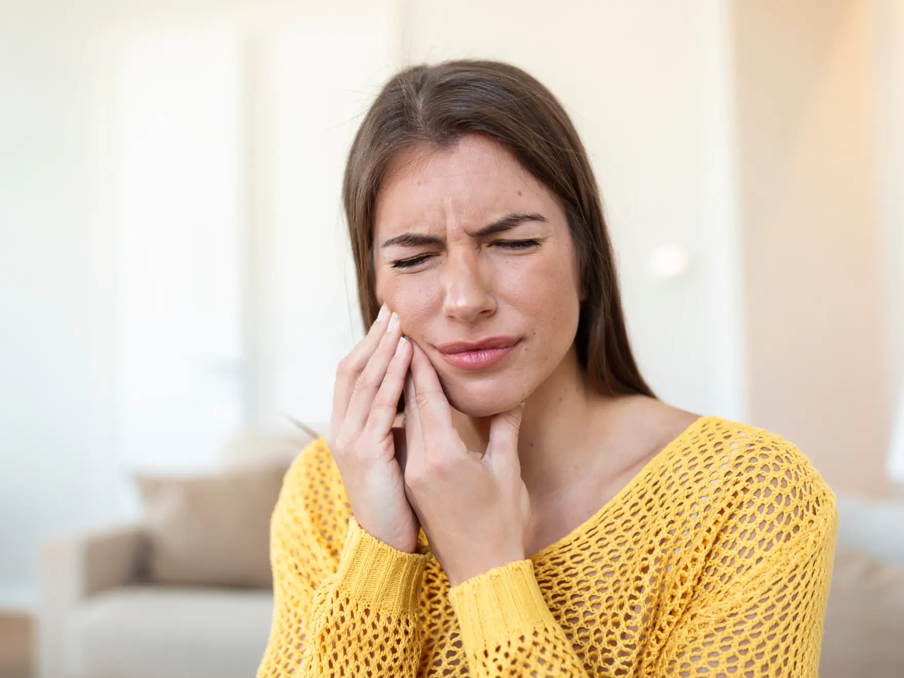 Woman in a yellow sweater holding her jaw with a painful expression, indicating toothache.