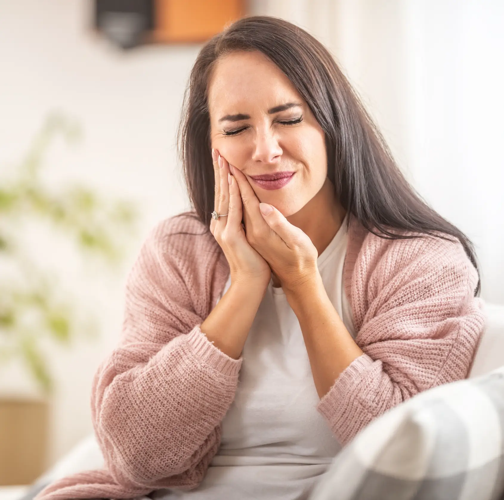 Woman with long brown hair wearing a pink sweater holding her cheek with a pained expression.