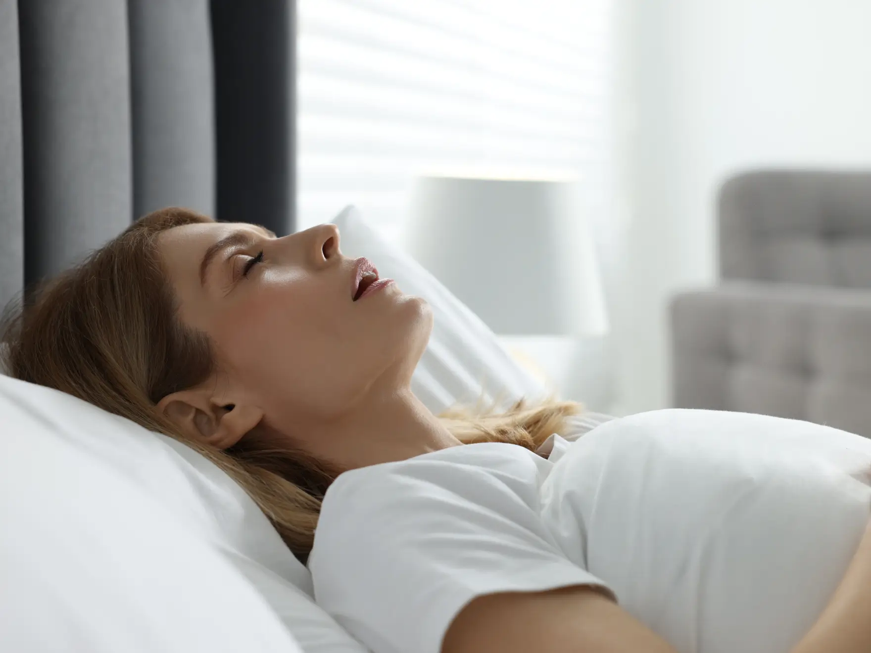 Woman sleeping on her back with mouth slightly open in a bright bedroom.