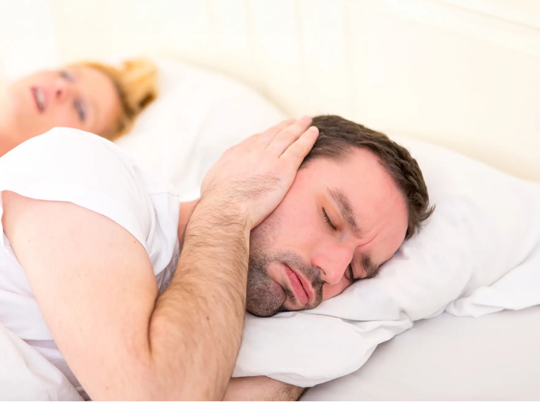 Man lying in bed with eyes closed covering his ear while a woman lies awake in the background.