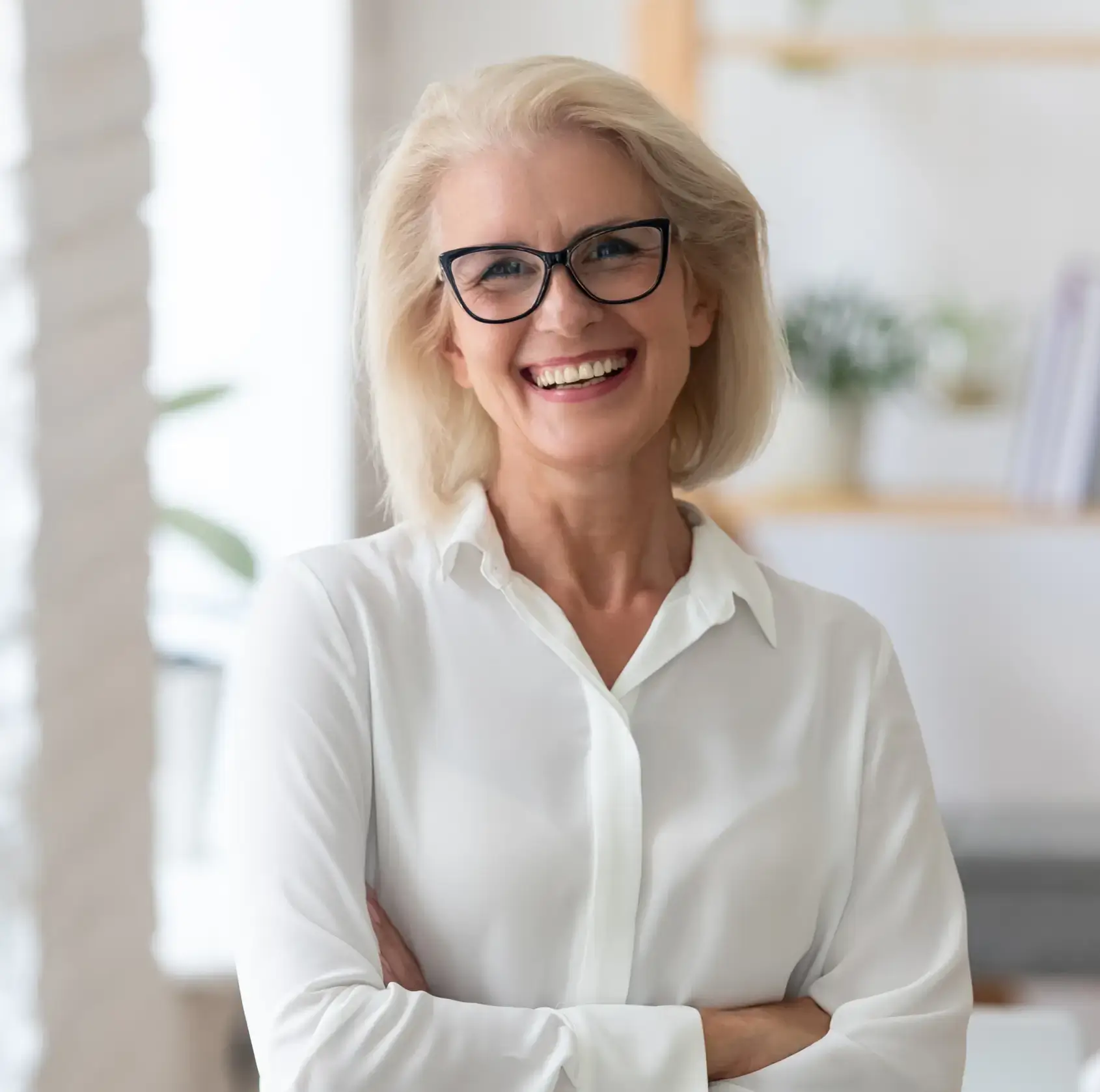 Smiling mature woman with short white hair and glasses wearing a white blouse, standing with arms crossed.