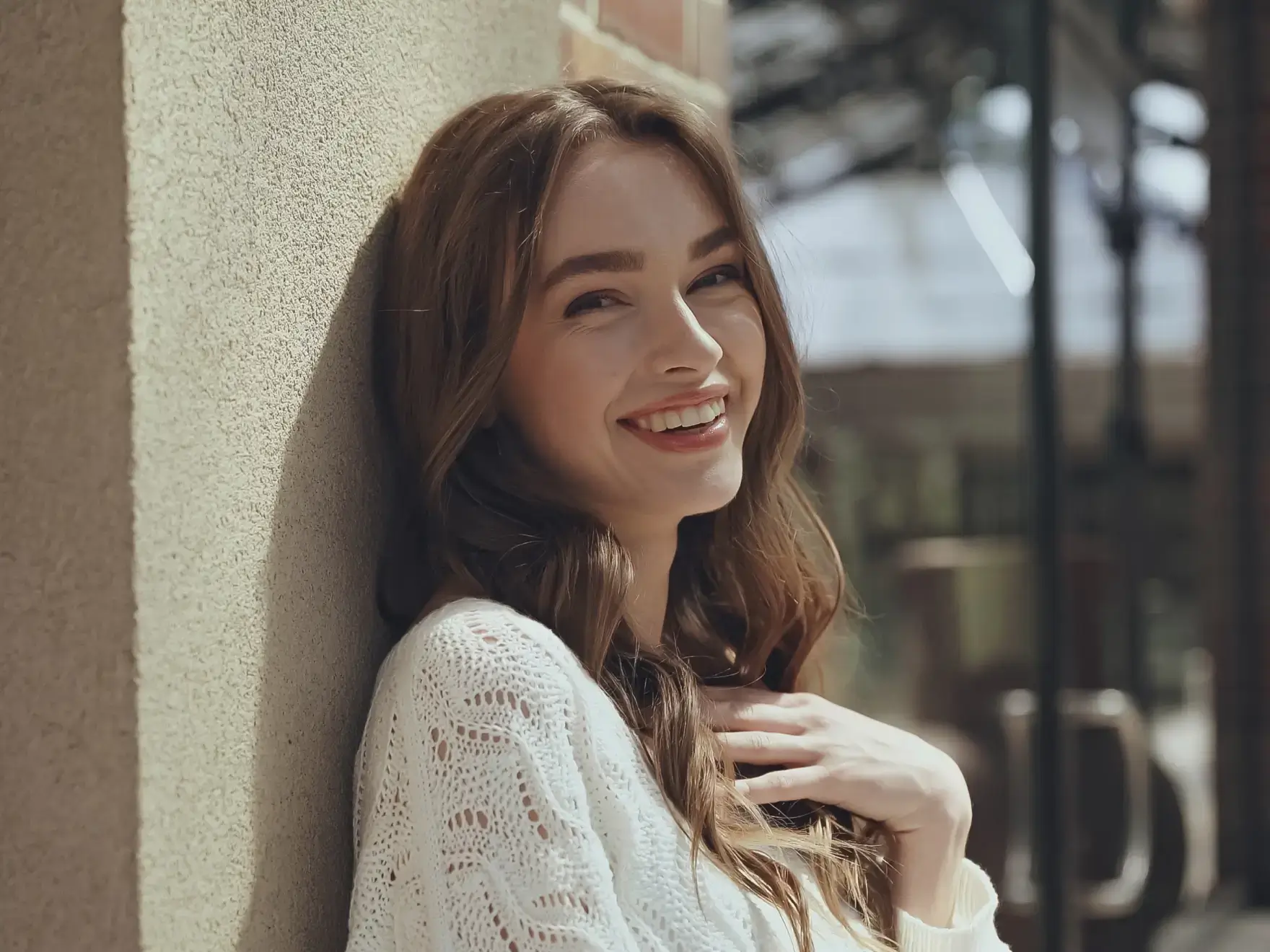 Young woman with long brown hair wearing a white sweater smiling while leaning against a textured wall outdoors.