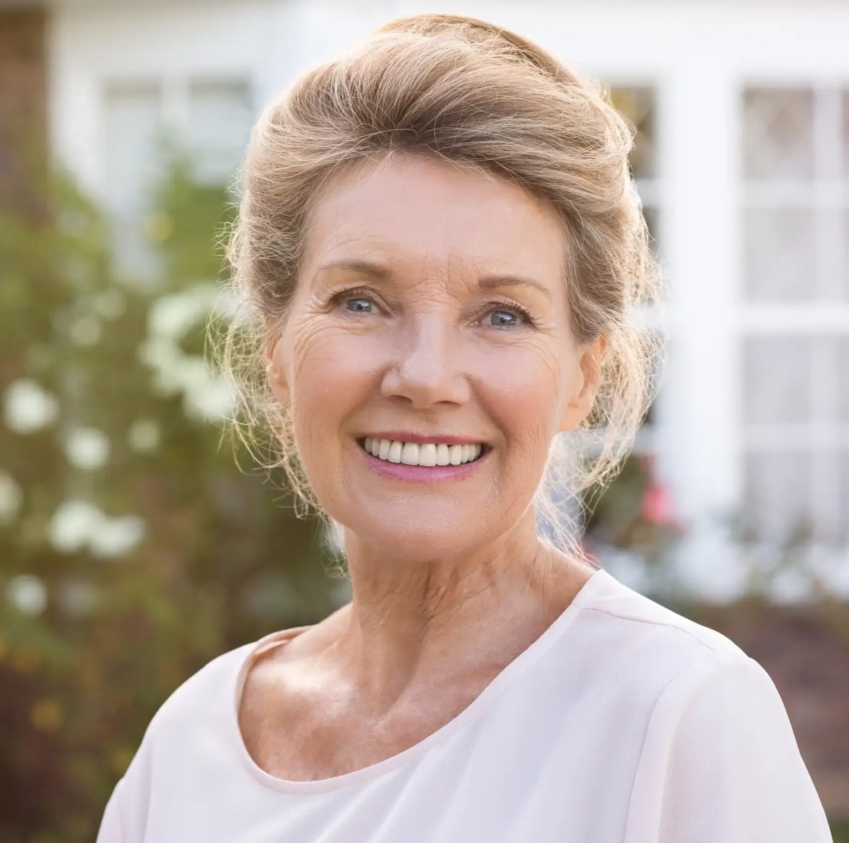 Smiling middle-aged woman with light brown hair and blue eyes wearing a light pink top outdoors.