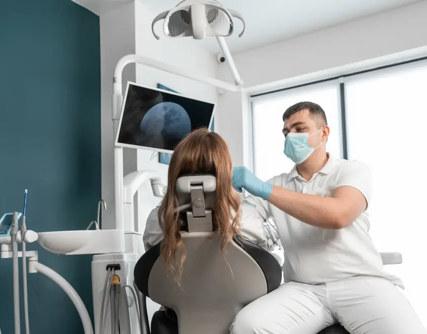 Dentist wearing mask and gloves examining a patient sitting in a dental chair in a modern clinic.