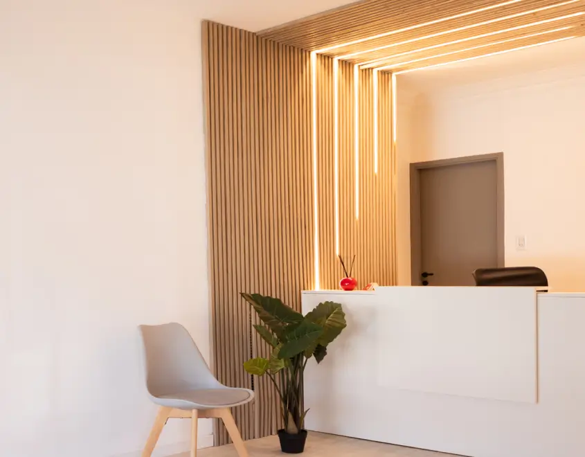 Minimalist reception area with a white desk, gray chair, green potted plant, and vertical wooden slats with integrated lighting on the wall.