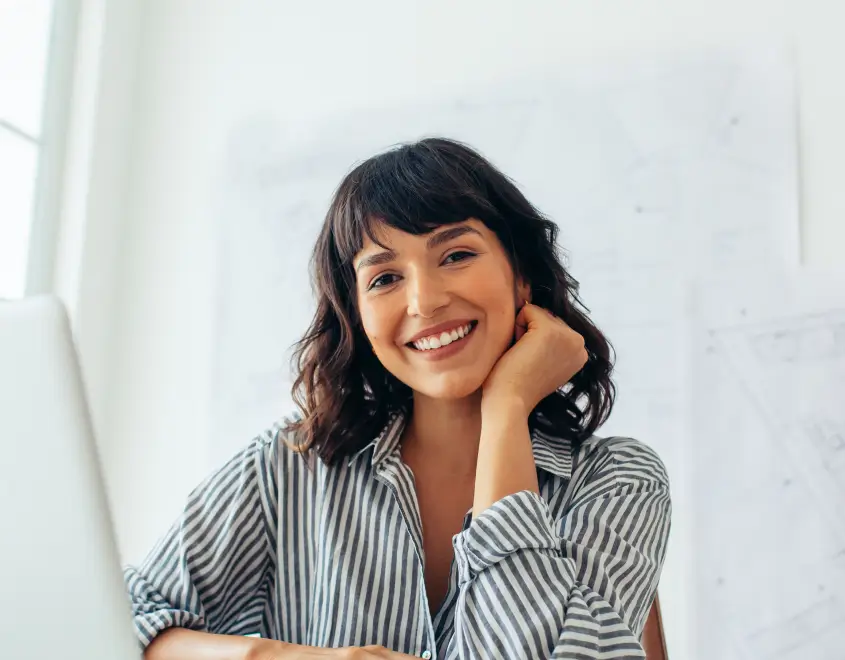 Smiling woman with dark curly hair wearing a striped shirt, sitting indoors near a window.