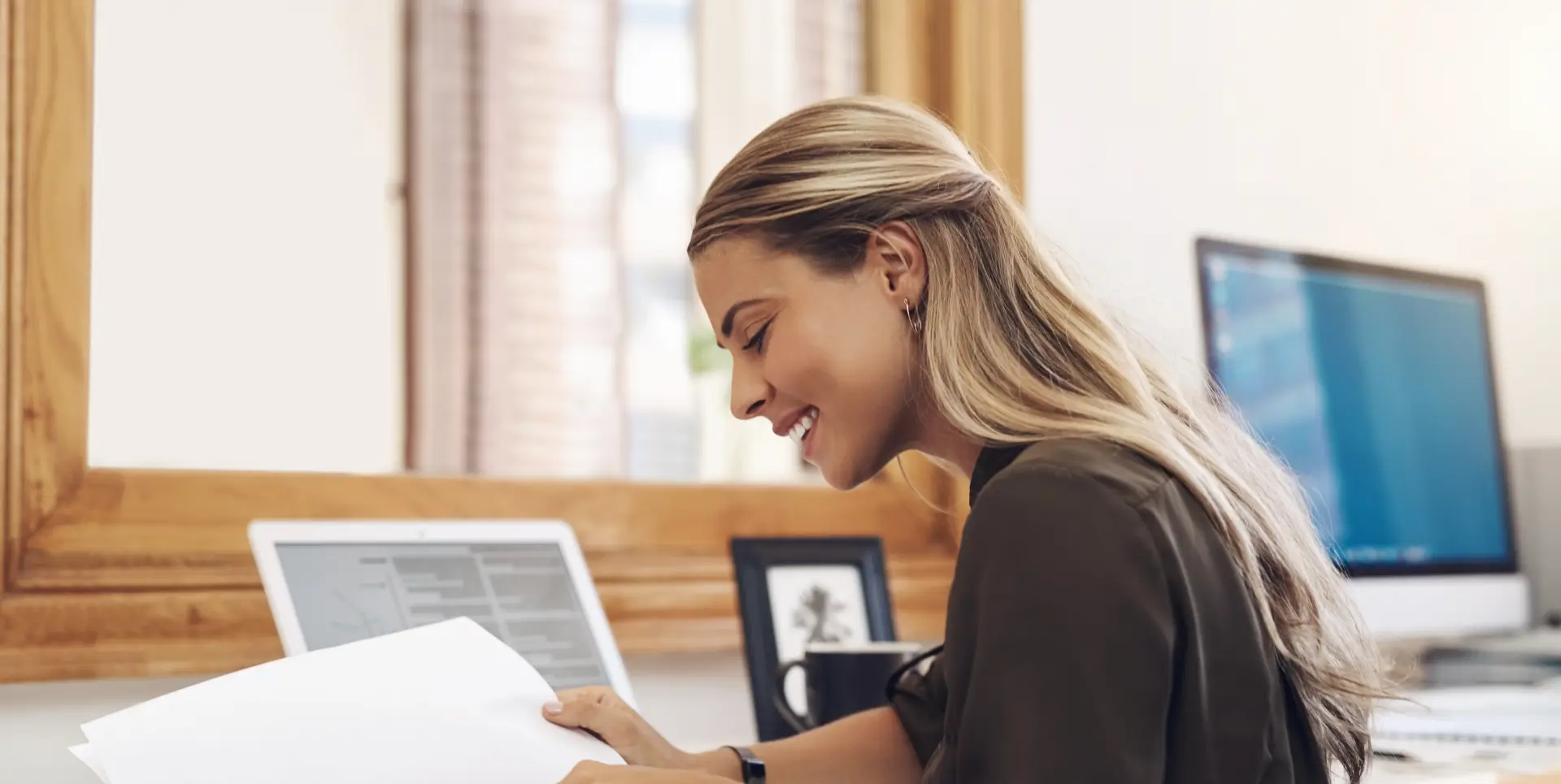 Smiling woman with blonde hair reading documents at a desk with laptop and desktop computer.