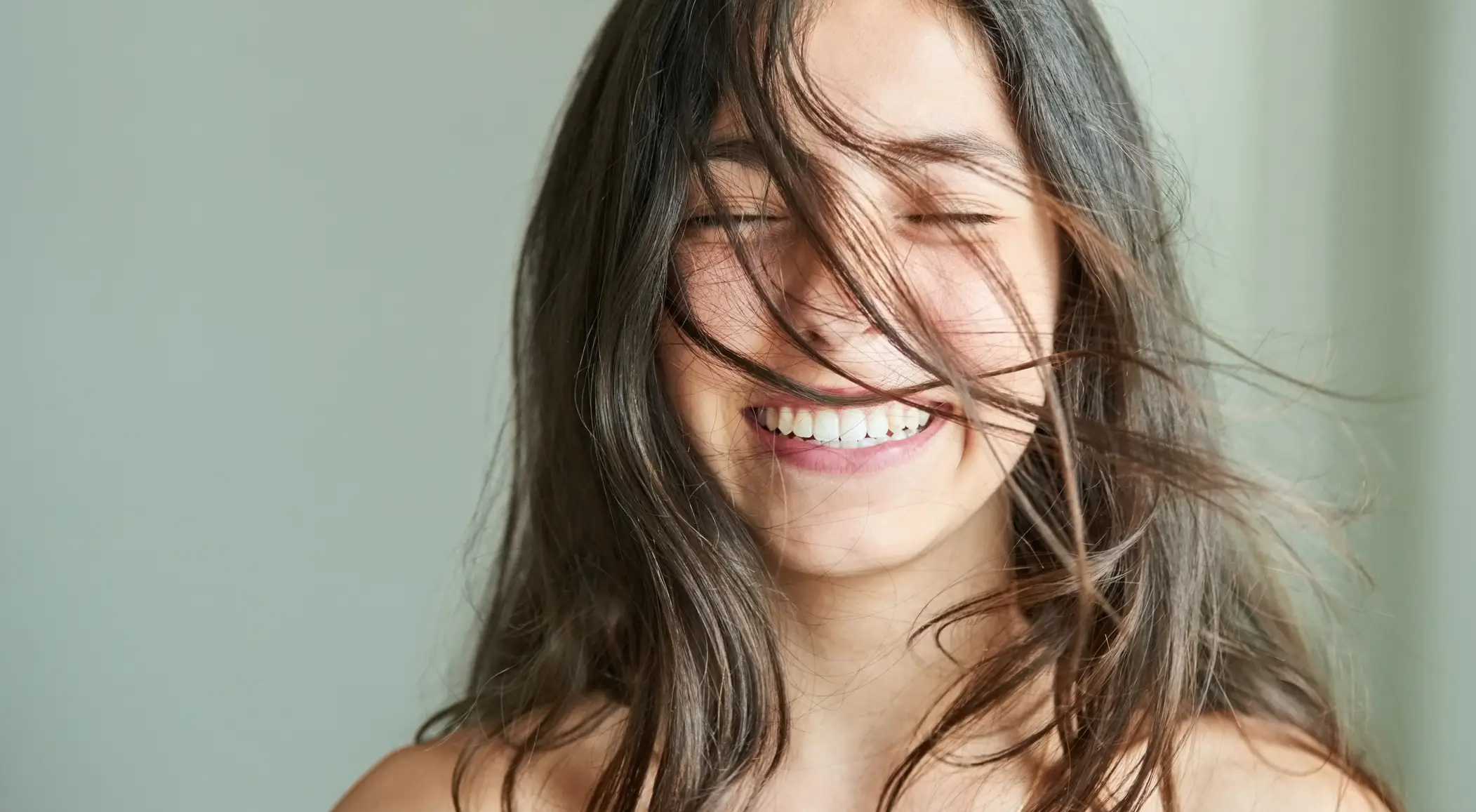 Close-up of a smiling woman with long hair blowing across her face and eyes closed.