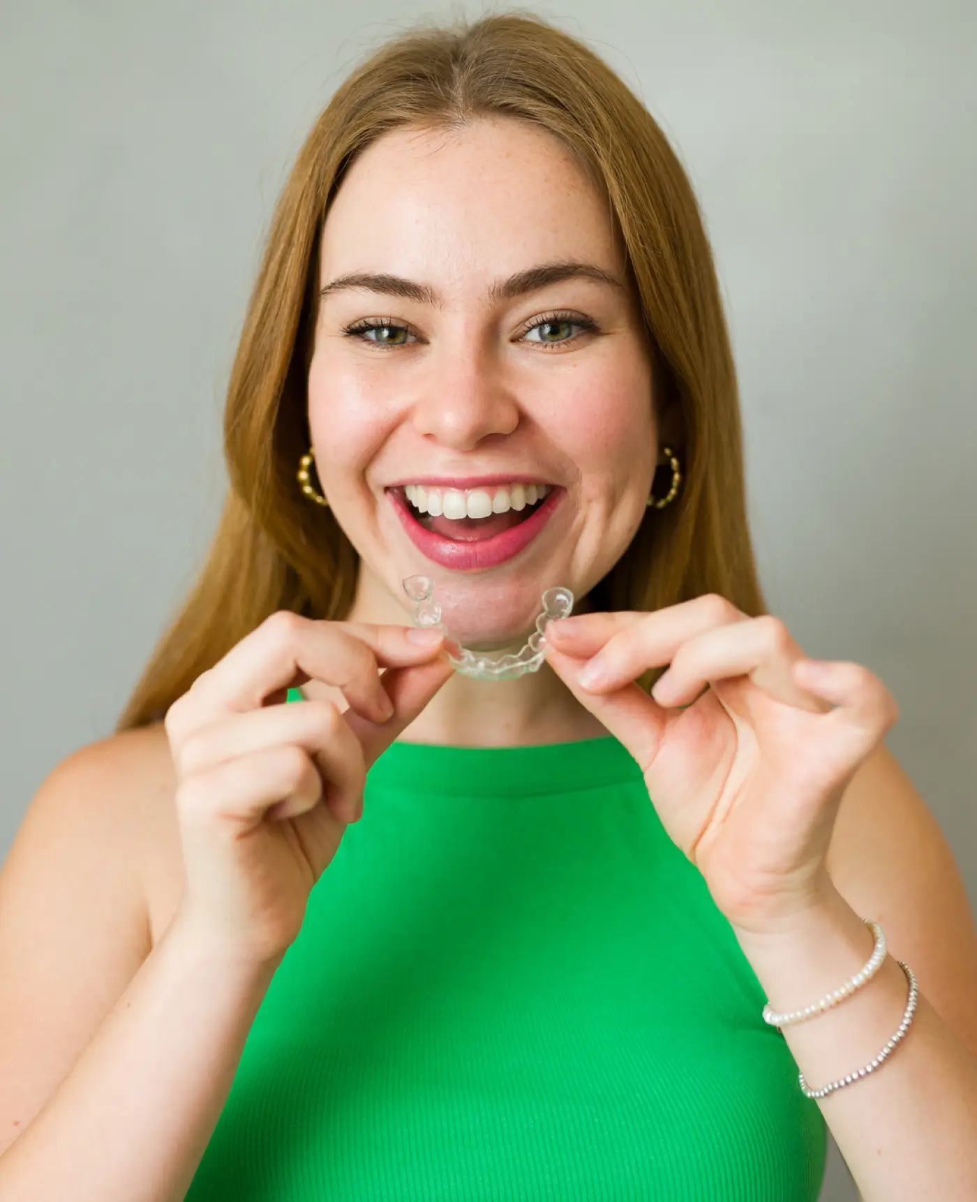 Smiling woman with long light brown hair holding a clear dental aligner in front of her mouth.