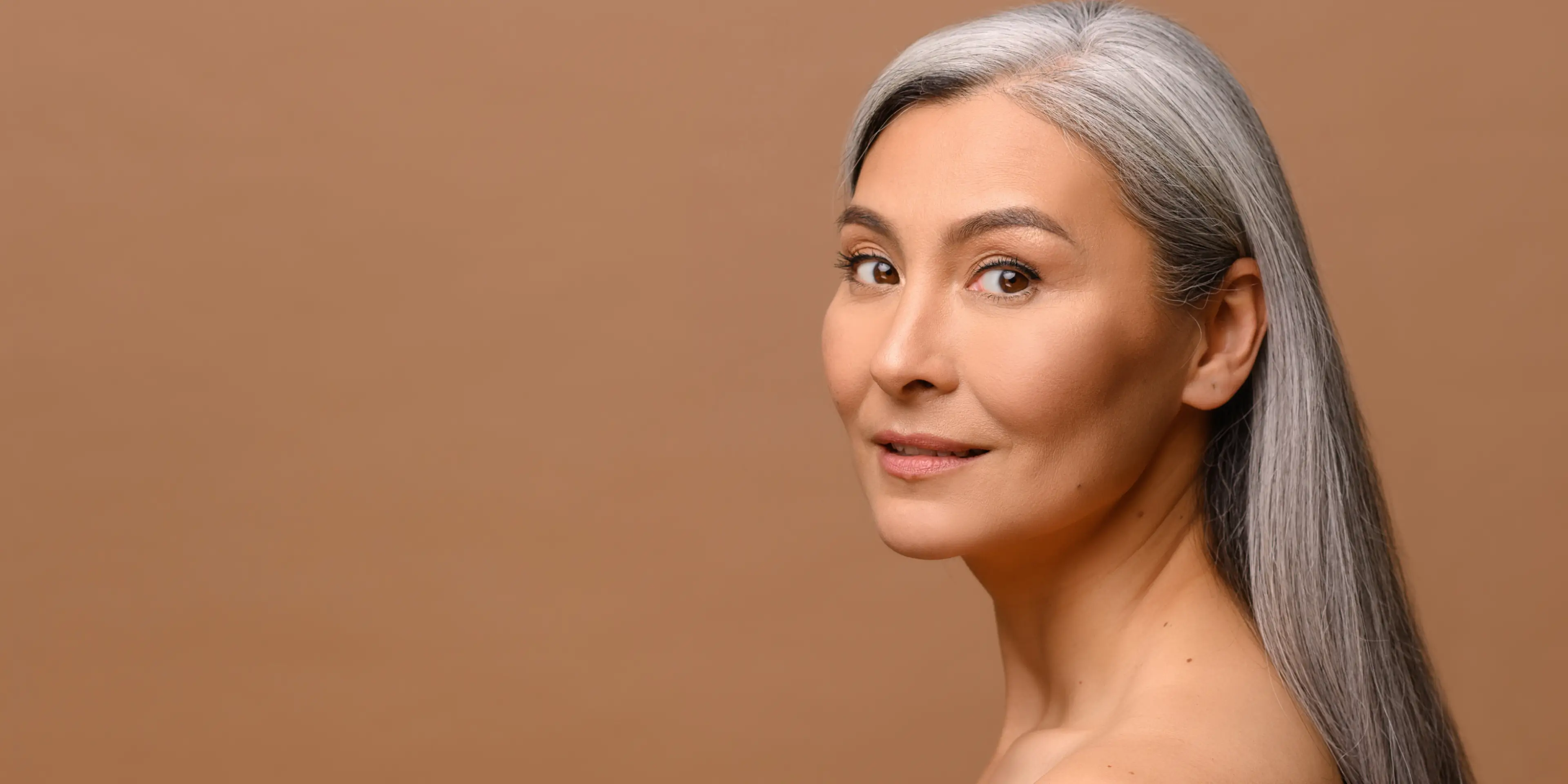 Portrait of a middle-aged woman with long gray hair looking over her shoulder against a brown background.
