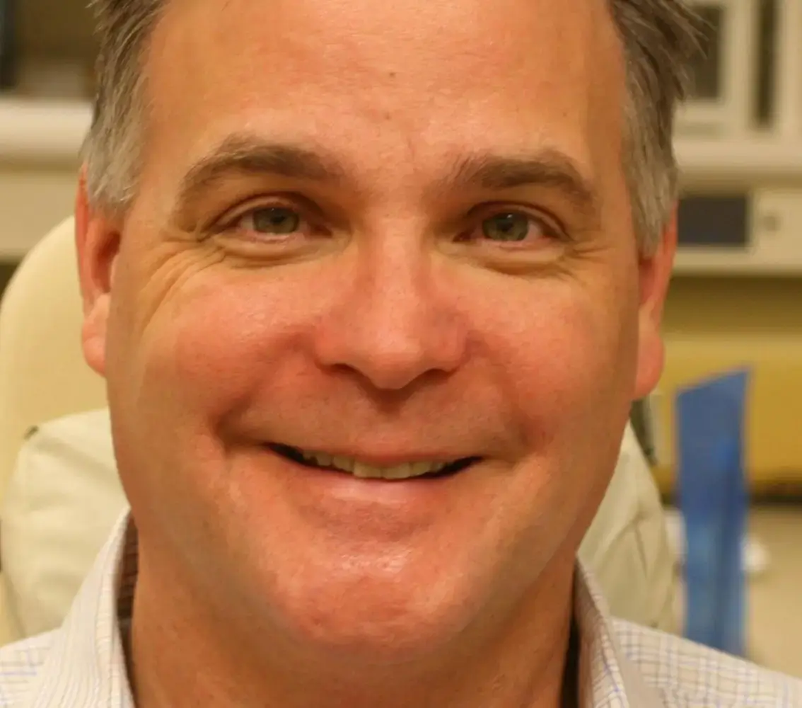Close-up of a middle-aged man smiling, with short graying hair and wearing a light-colored shirt.